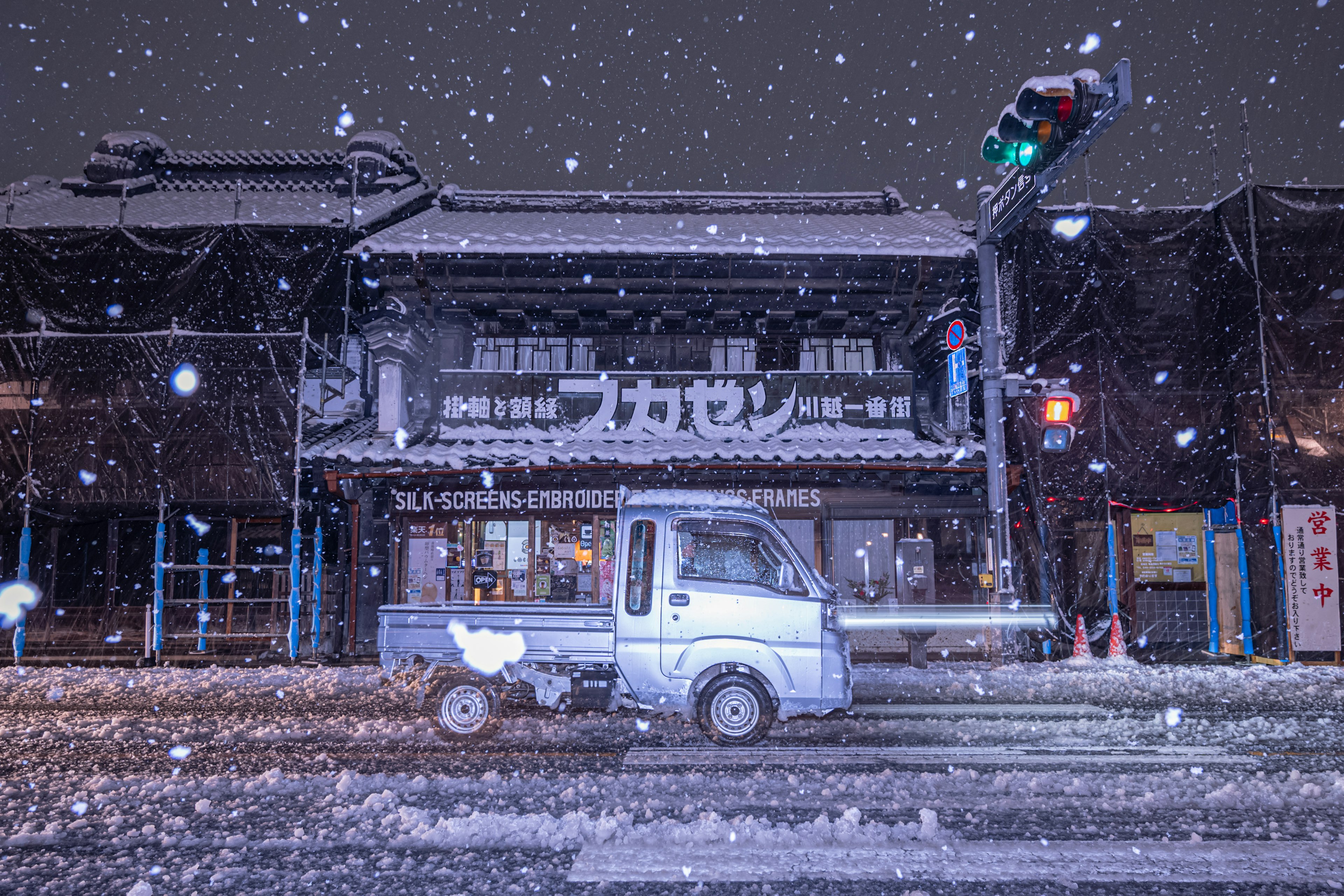 雪の中を走る小型トラックと伝統的な建物が映る夜の風景