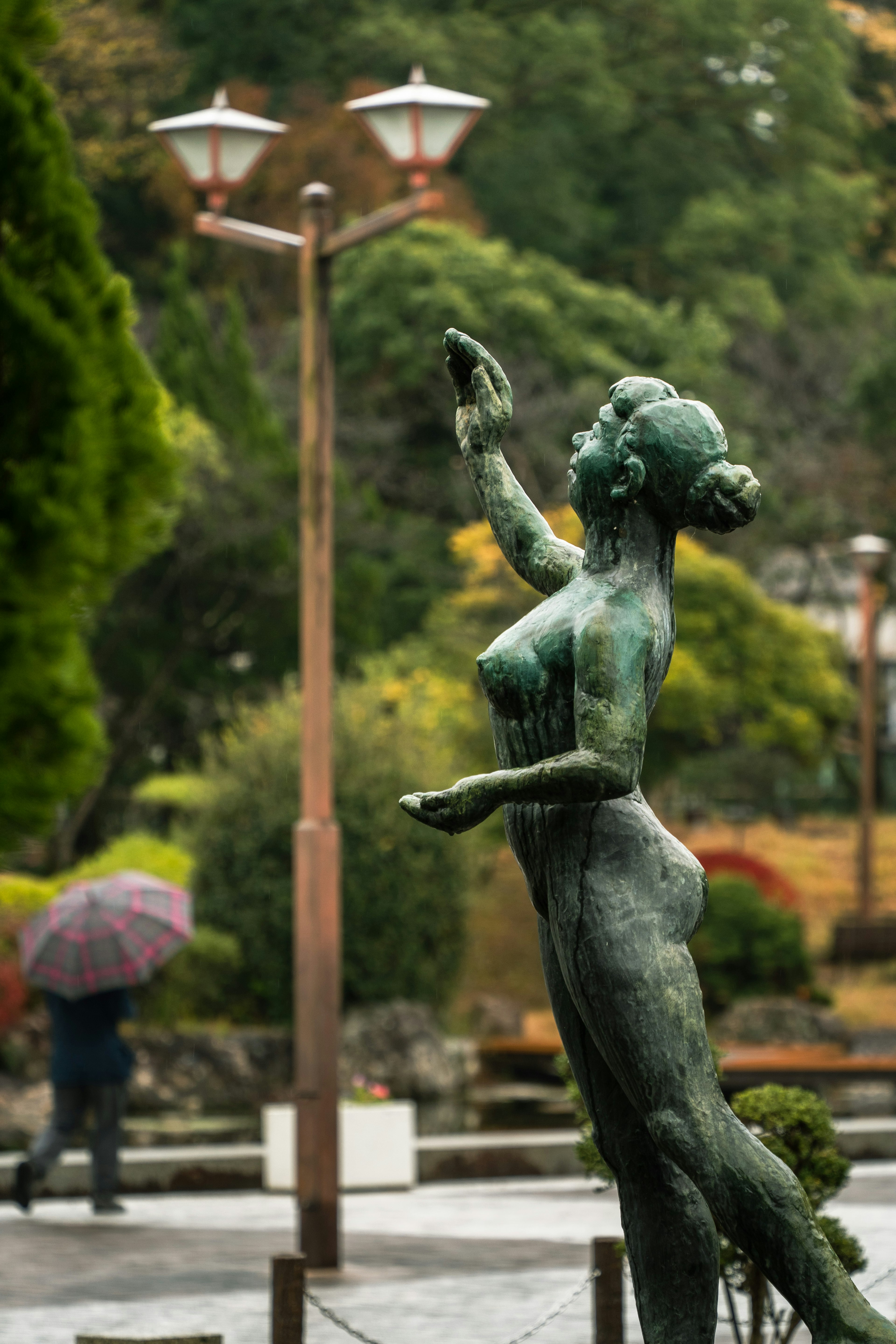 公園で立っている青銅の女性像　周囲に木々と街灯　背後に傘を持った人