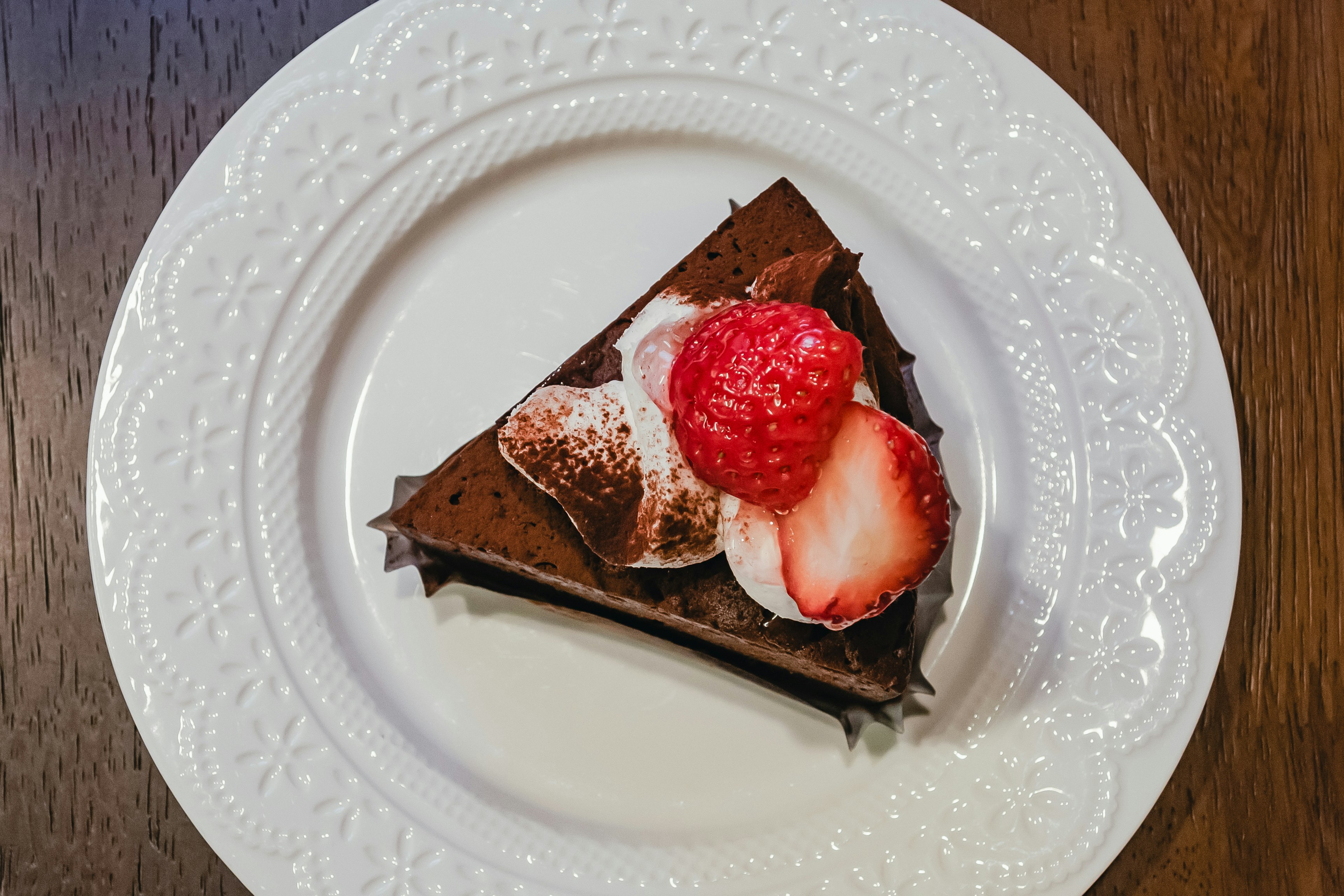 Seiris kue cokelat dihiasi dengan stroberi segar di atas piring putih yang elegan