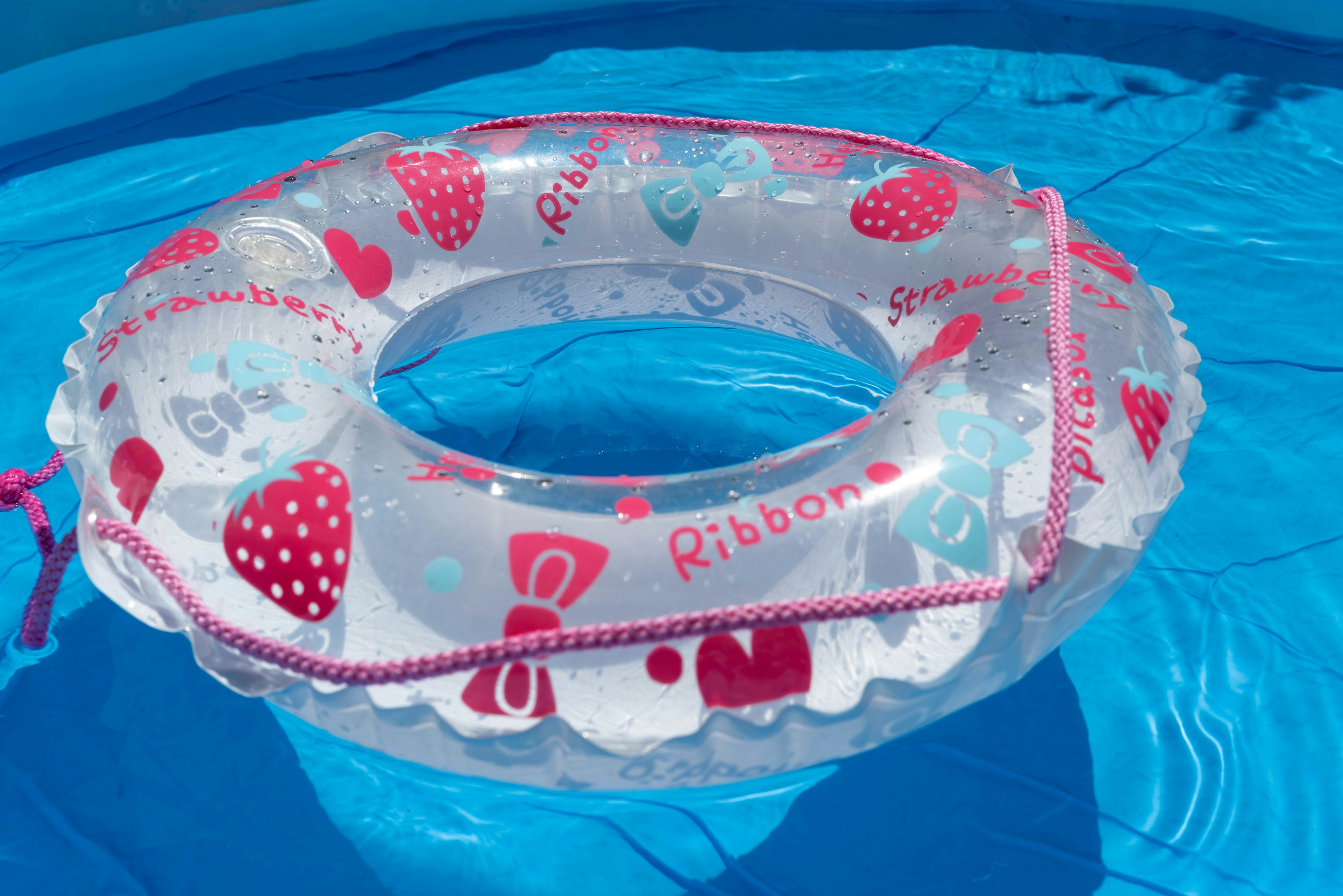 Transparent inflatable ring with red strawberries and ribbon design floating in blue pool water