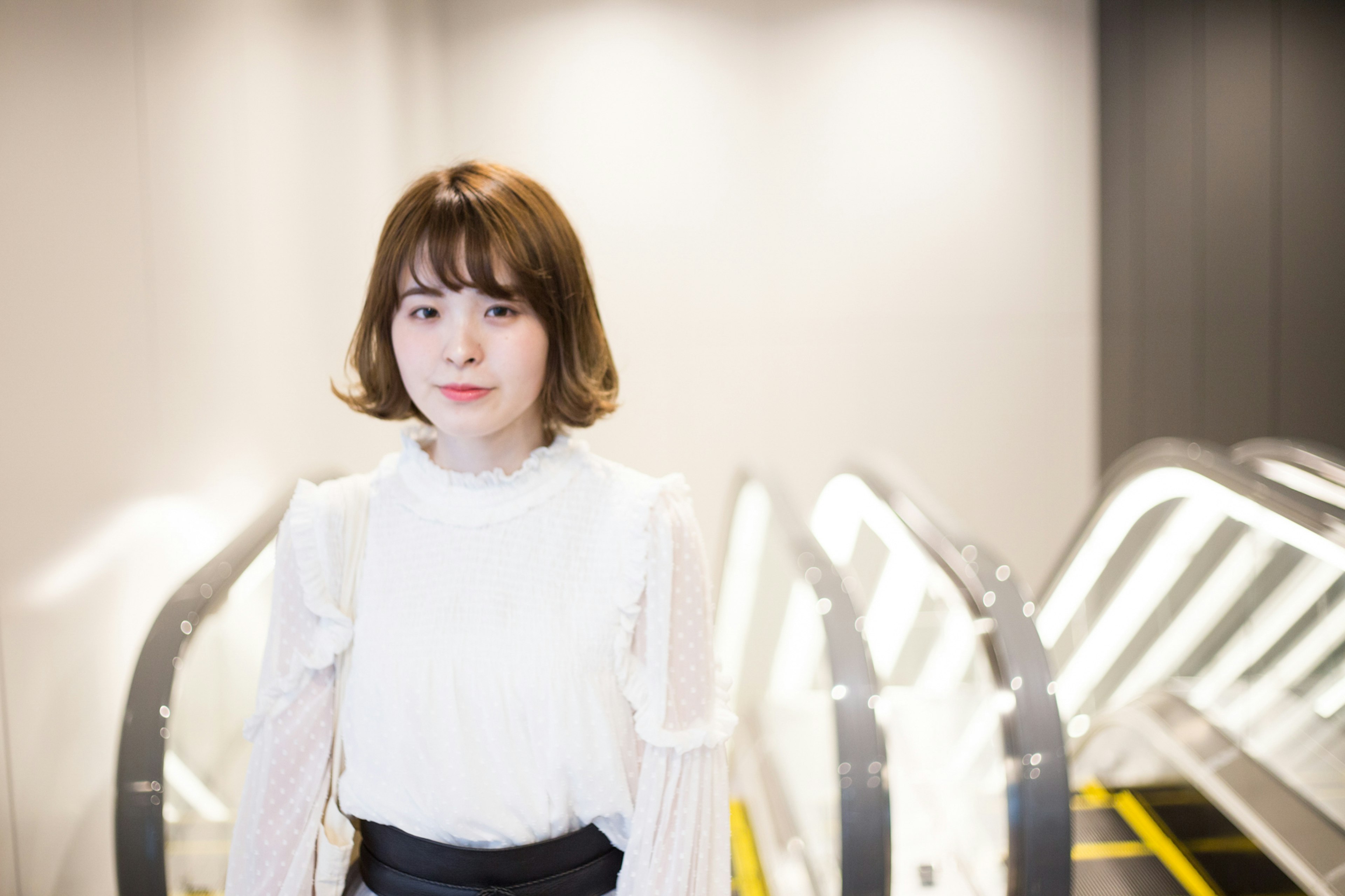 A young woman wearing a white blouse stands against a bright escalator background