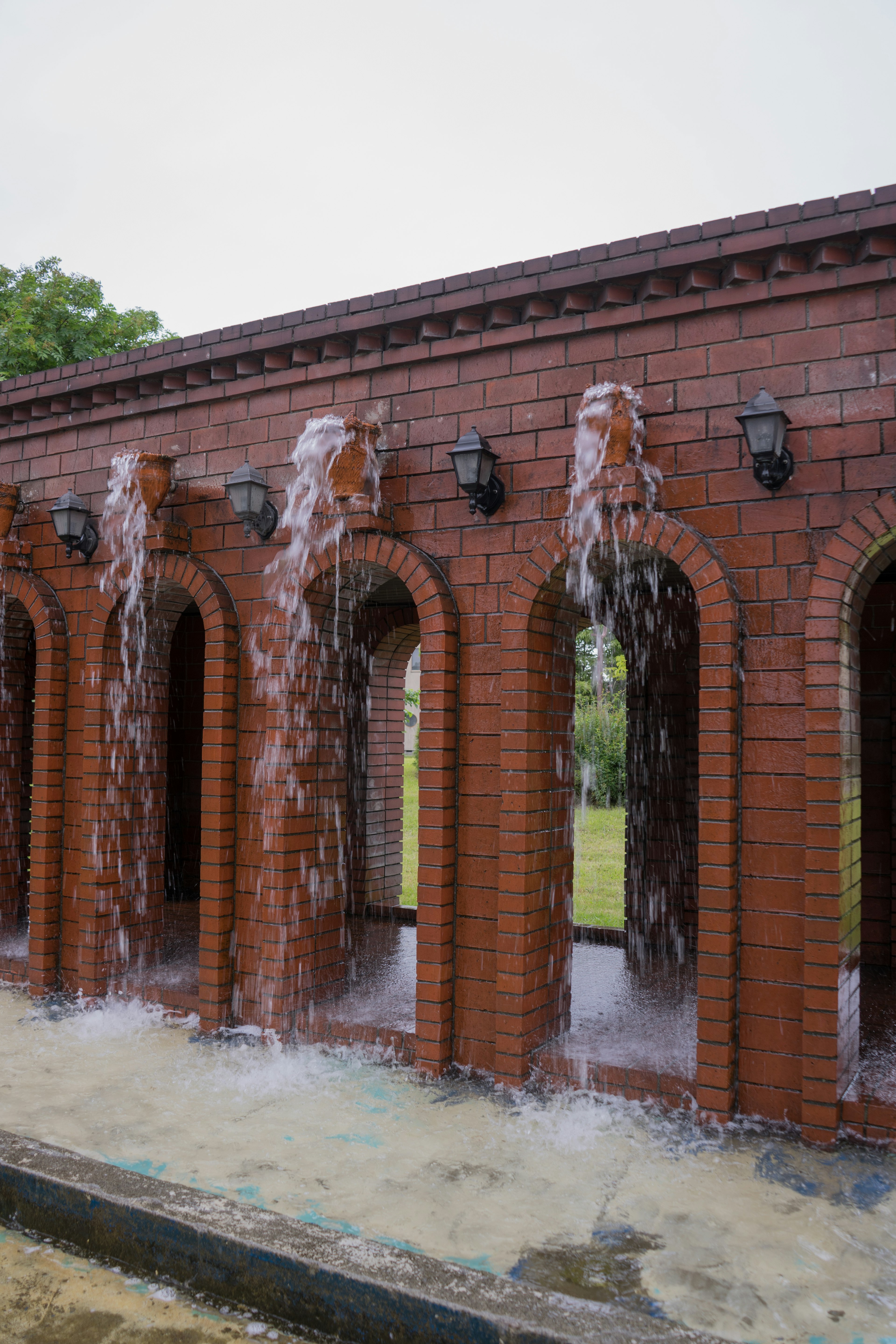 赤茶色の壁に水が流れるアーチ型の噴水が特徴的な公園の景色