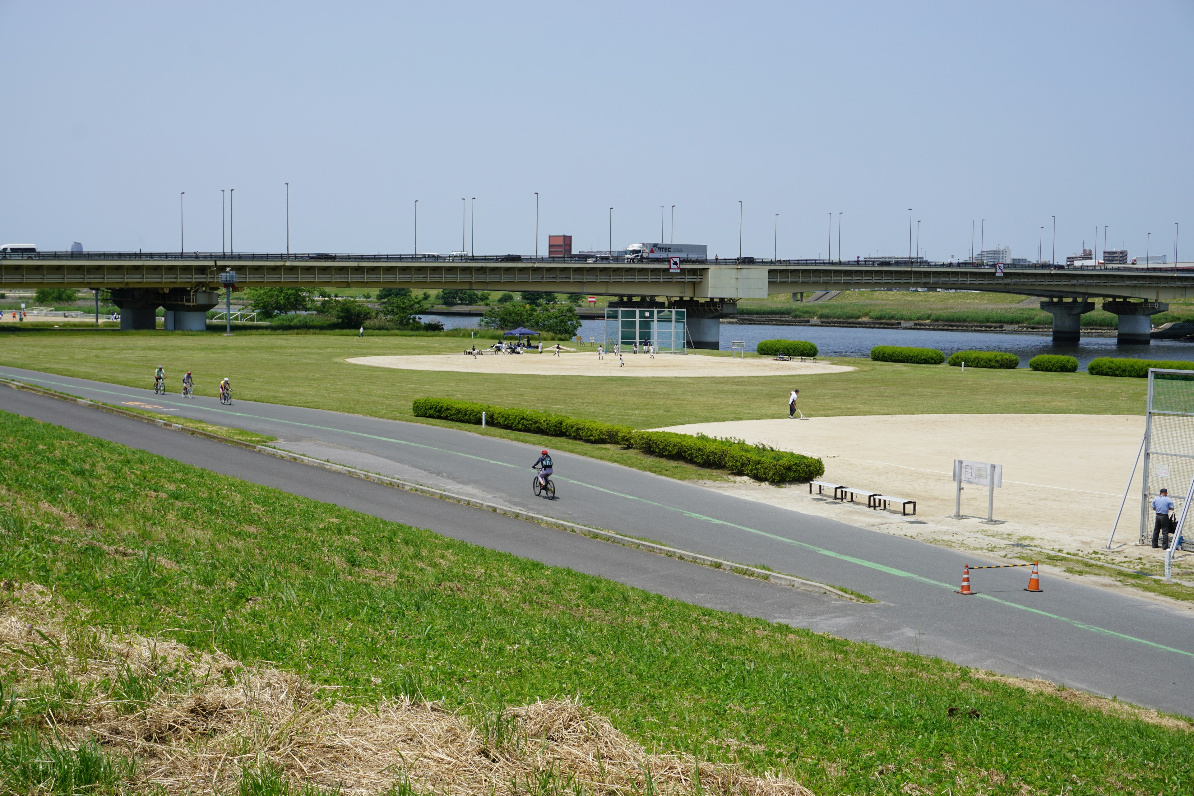 青空の下で川沿いの道を自転車で走る人々と橋が見える風景