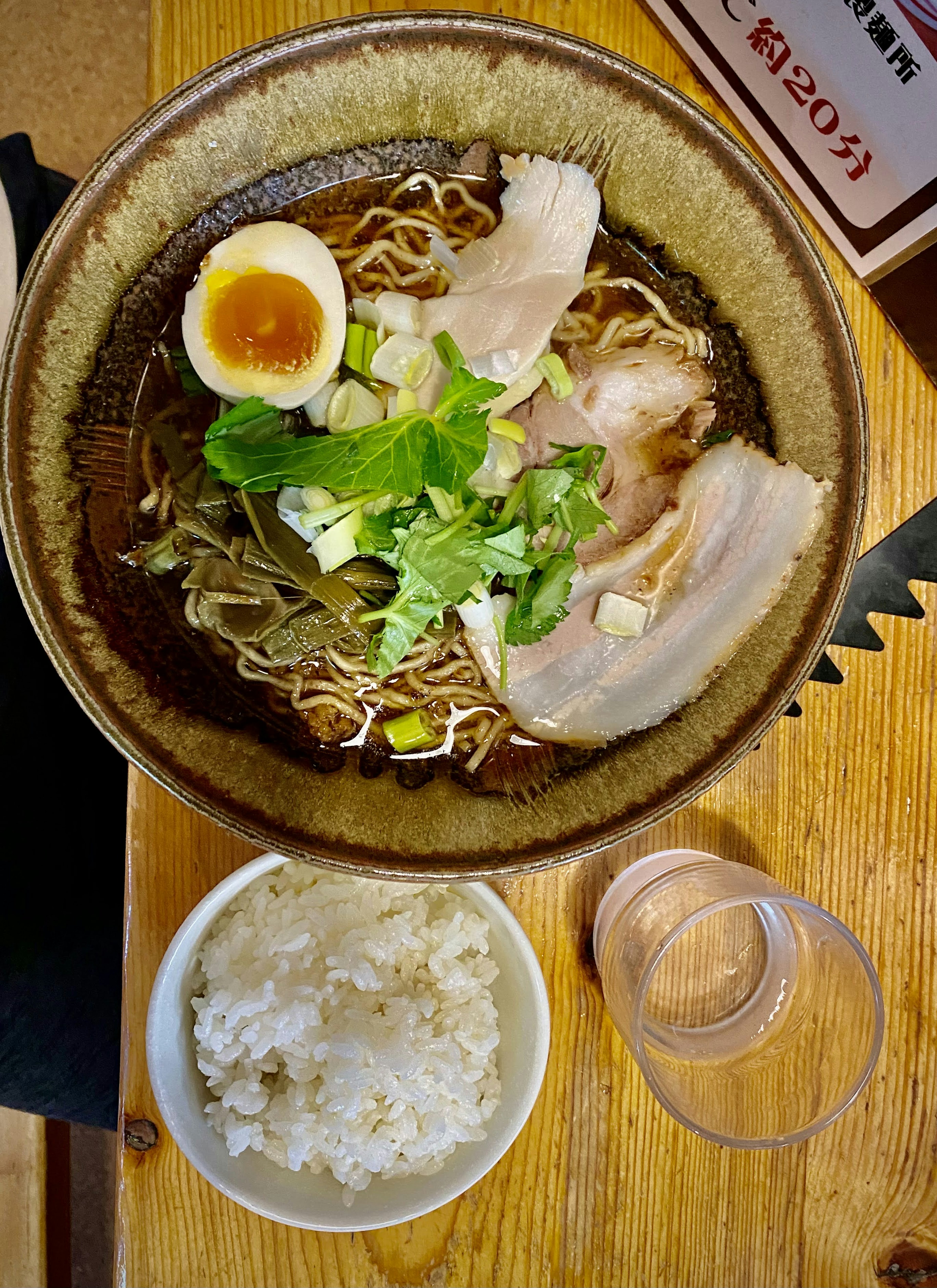 ラーメンのボウルにチャーシュー、煮卵、ネギ、香草が盛り付けられている