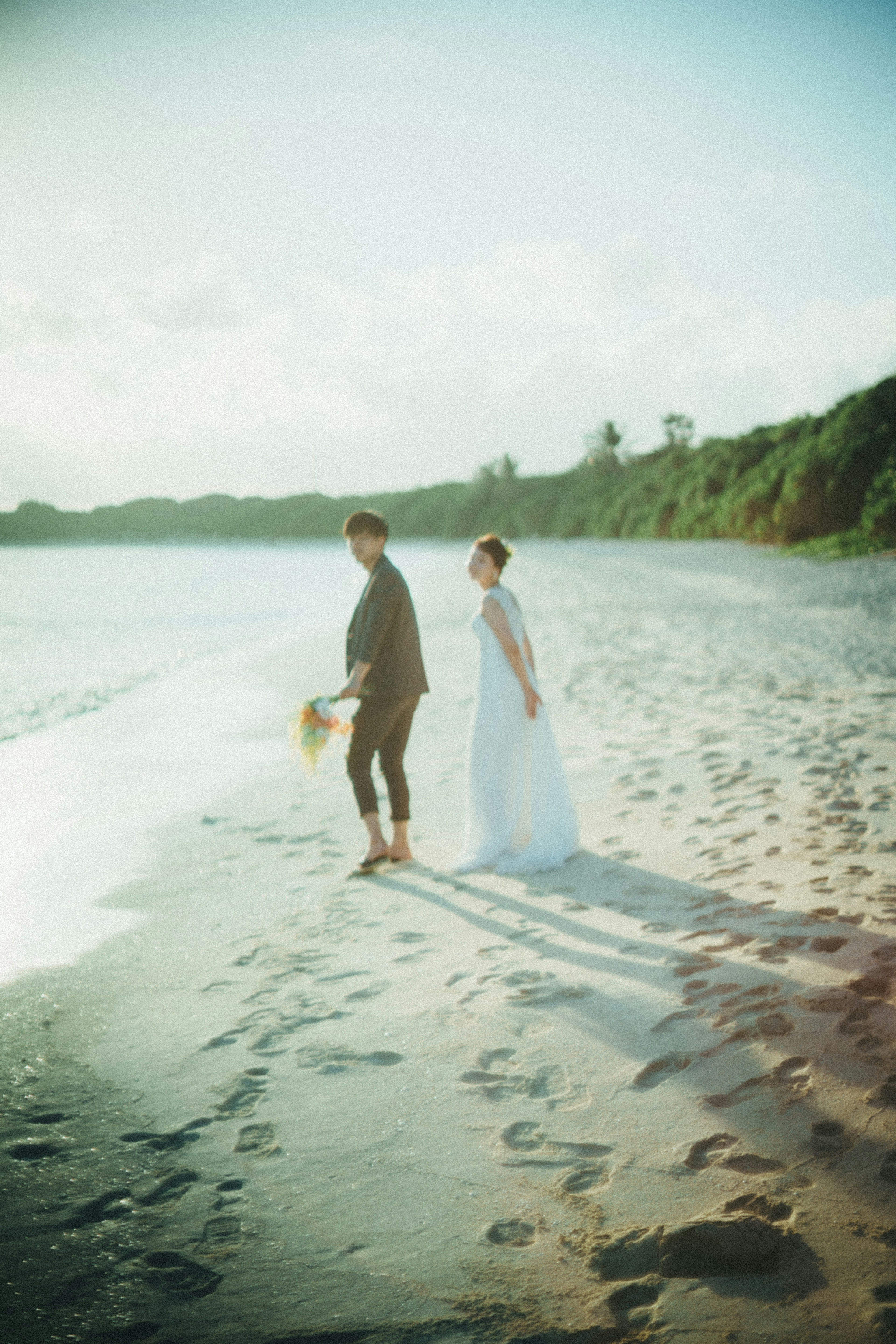 คู่รักยืนอยู่บนชายหาดจับมือกันในชุดเดรสสีขาว