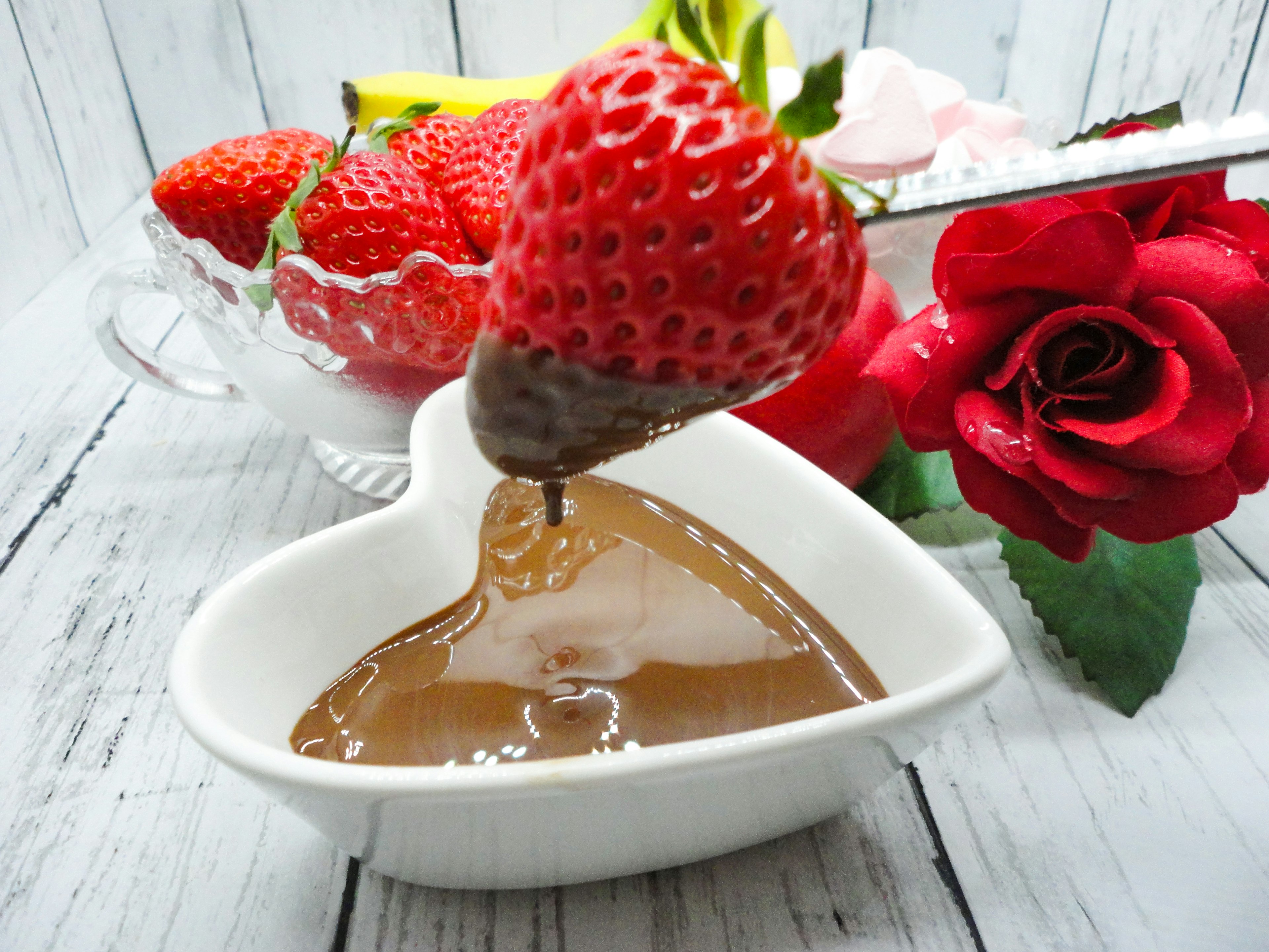 Fragola immersa in salsa di cioccolato su un piatto a forma di cuore con rose rosse e fragole sullo sfondo