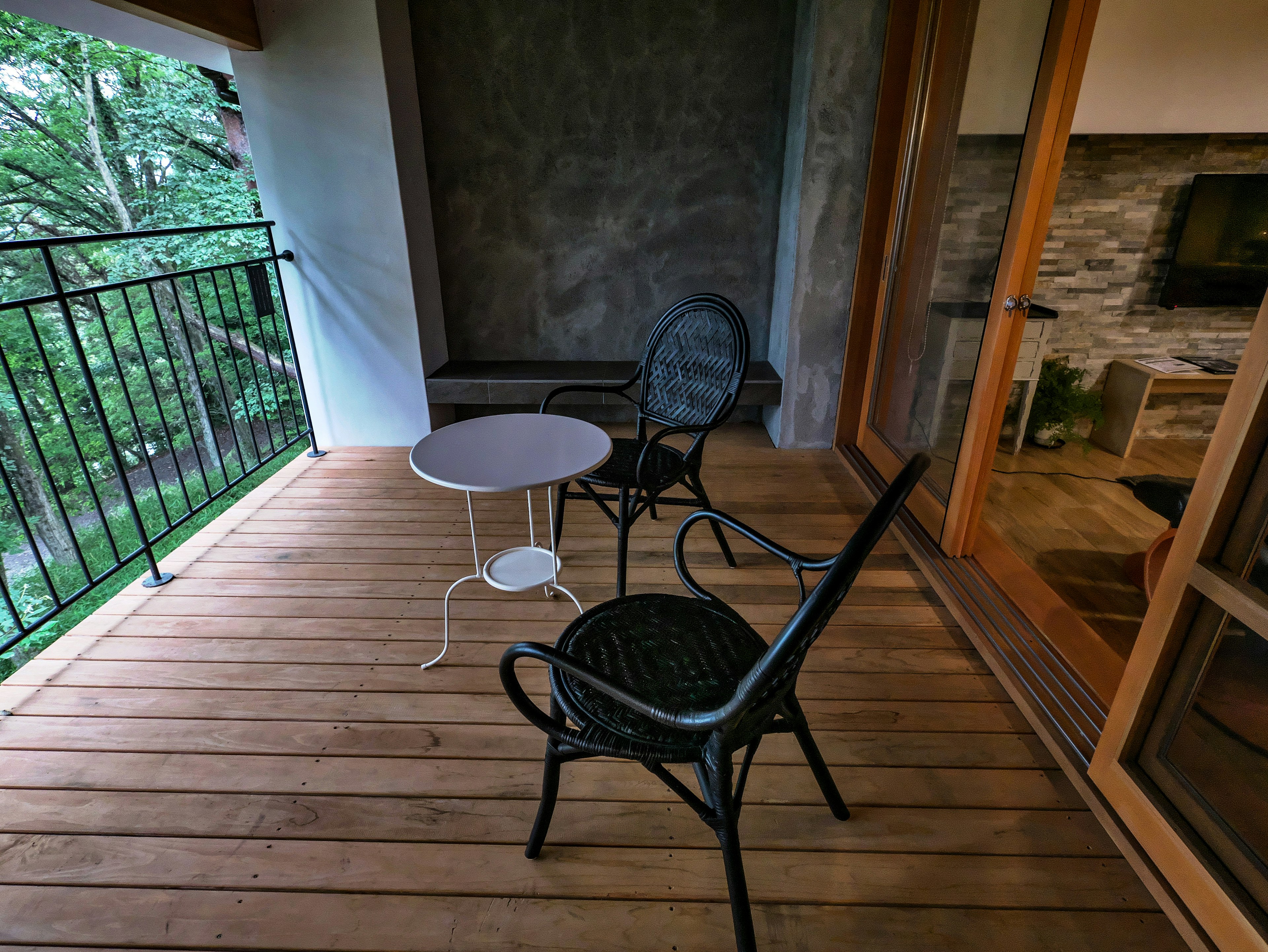 A balcony with a wooden deck featuring a white table and a black chair overlooking greenery