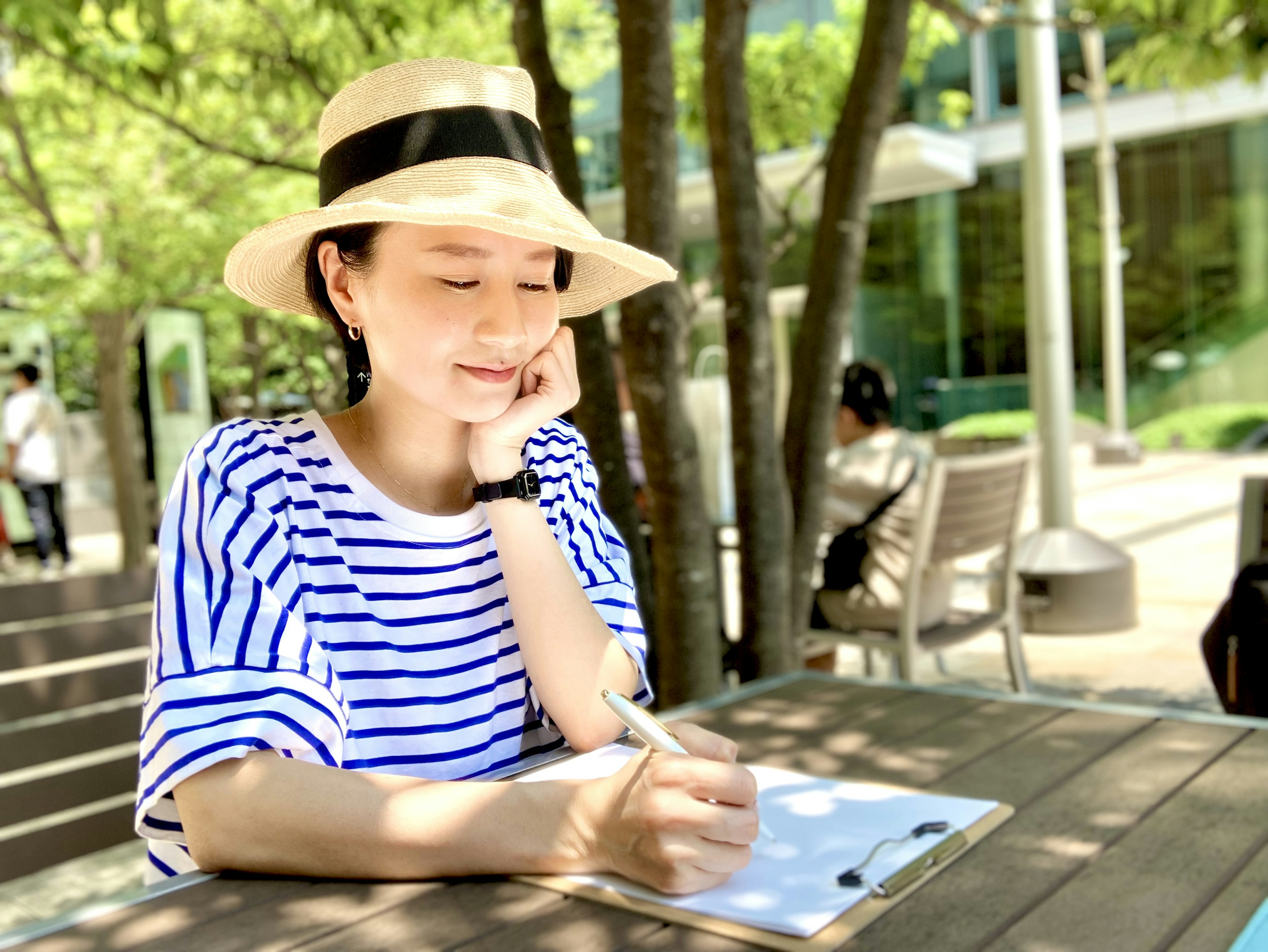 A woman in a striped shirt wearing a hat sitting under a tree writing in a notebook