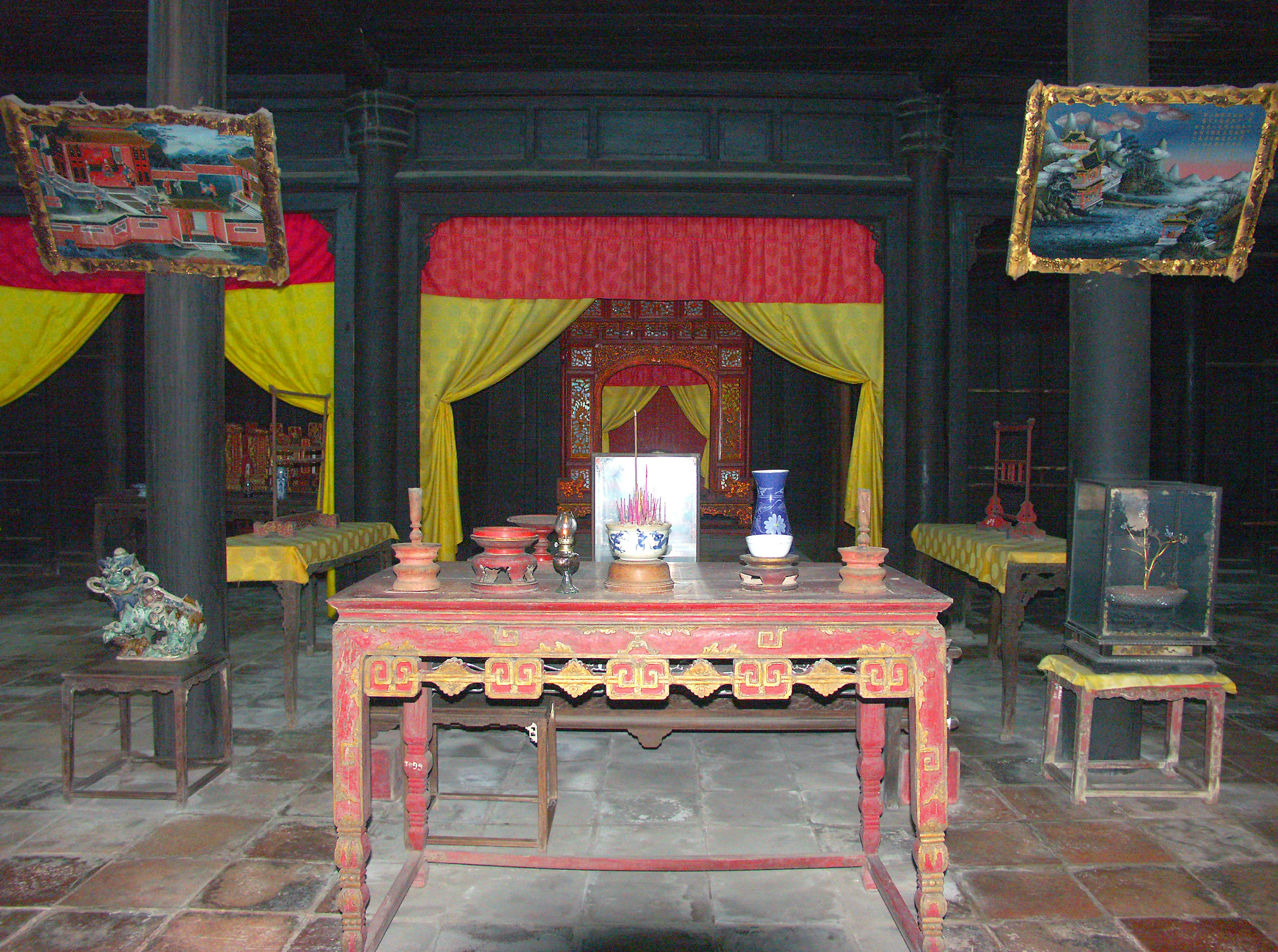 Interior tradicional con mesa de madera y sillas adornadas con elementos decorativos Cortinas rojas y amarillas al fondo Pinturas distintivas colgadas en las paredes