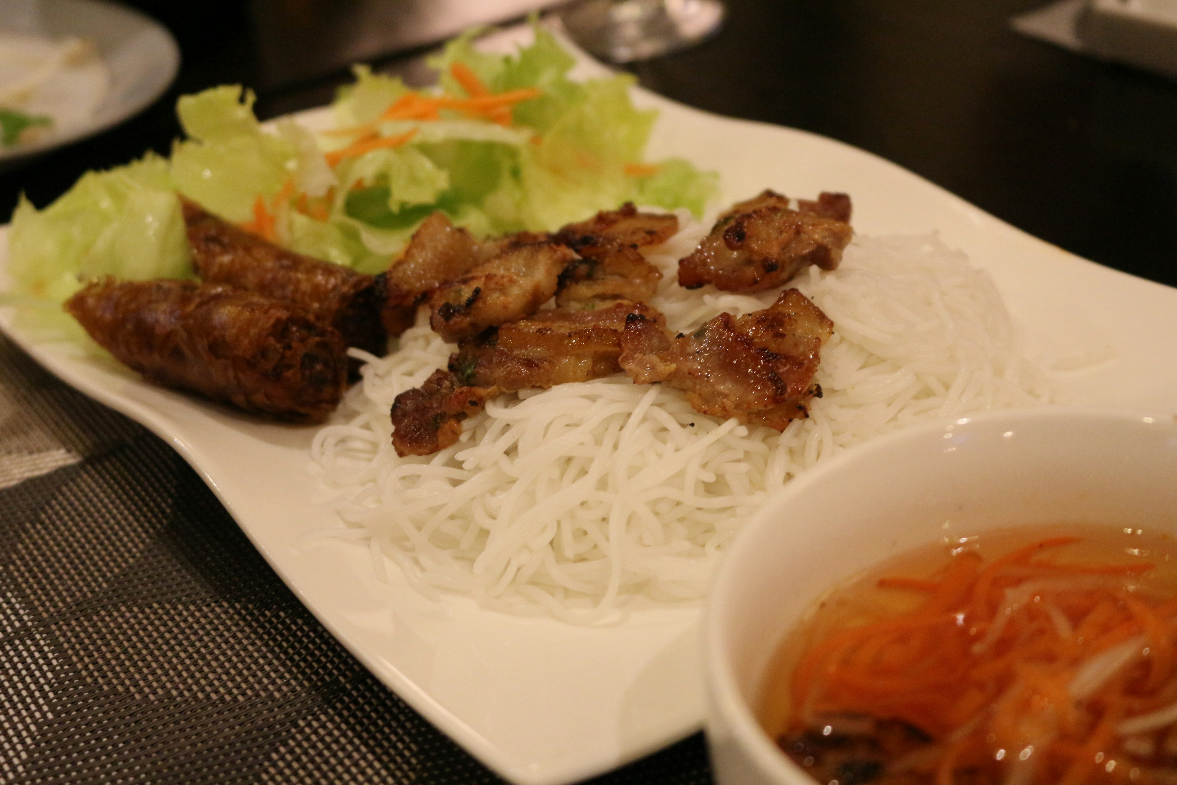 Dish of rice noodles topped with grilled meat fresh lettuce and salad on a white plate alongside dipping sauce