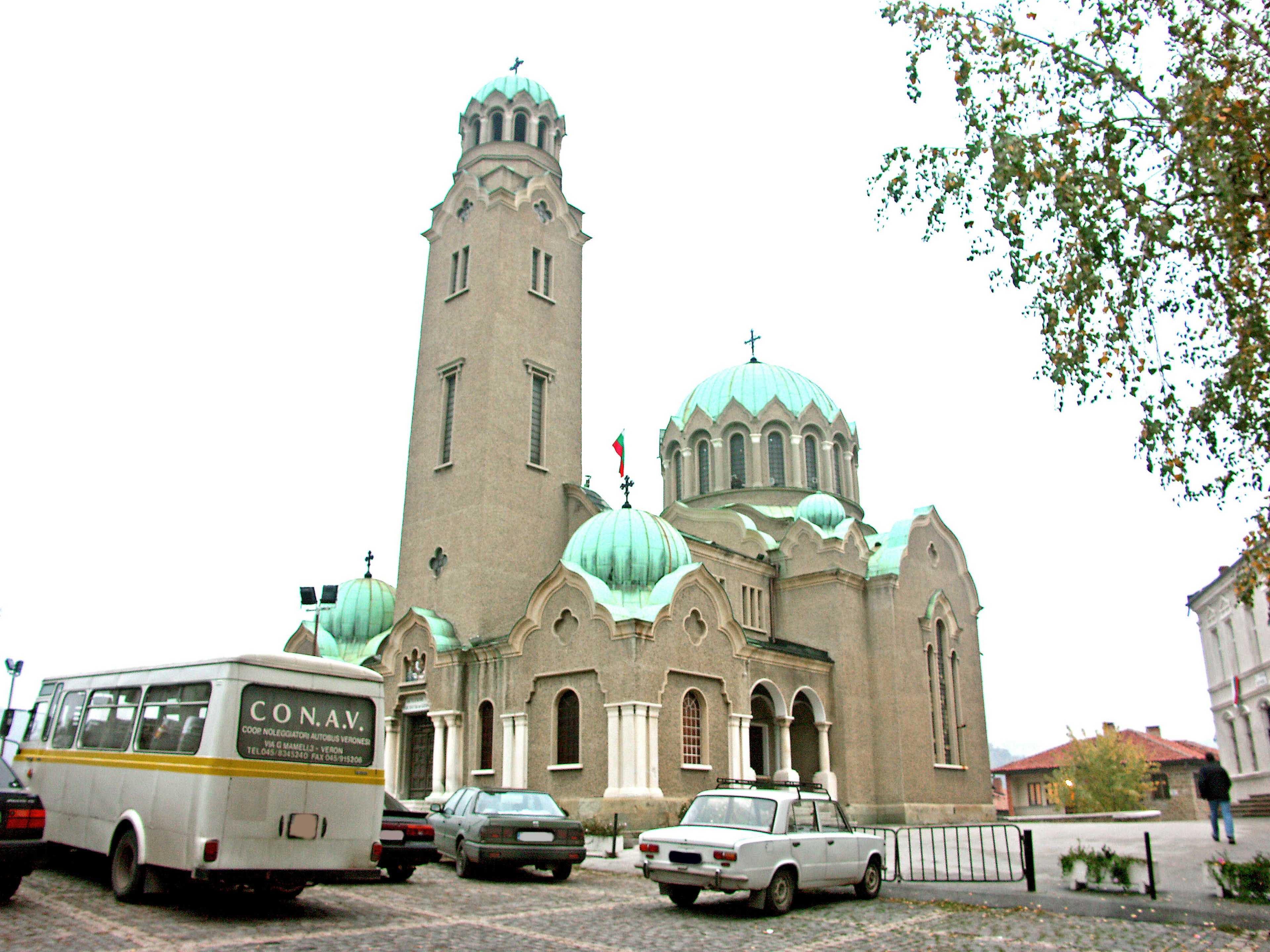 Church exterior with green domes and tall tower parked bus and cars nearby