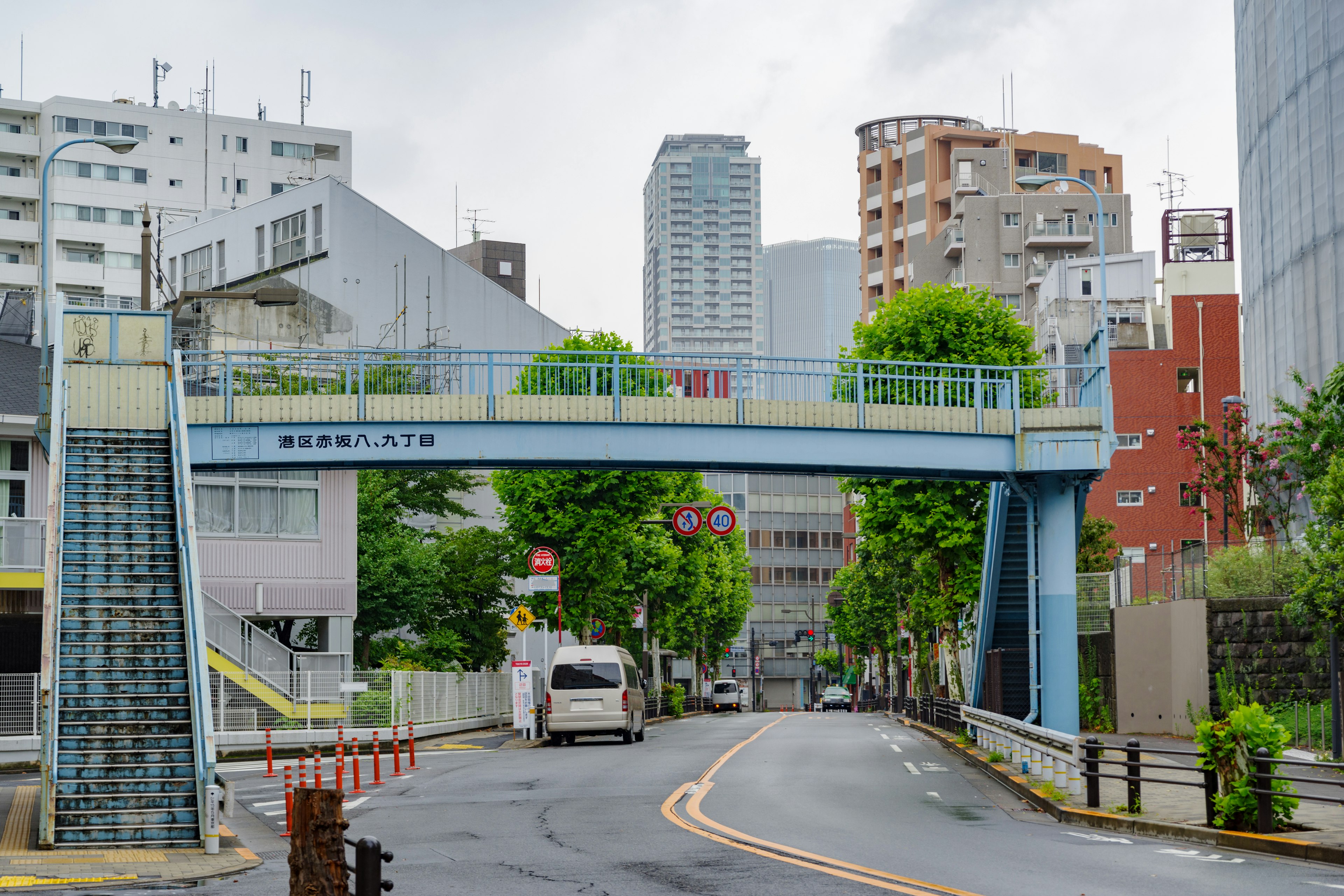 ภาพสะพานคนเดินสีน้ำเงินที่ล้อมรอบด้วยต้นไม้สีเขียวและทิวทัศน์เมือง