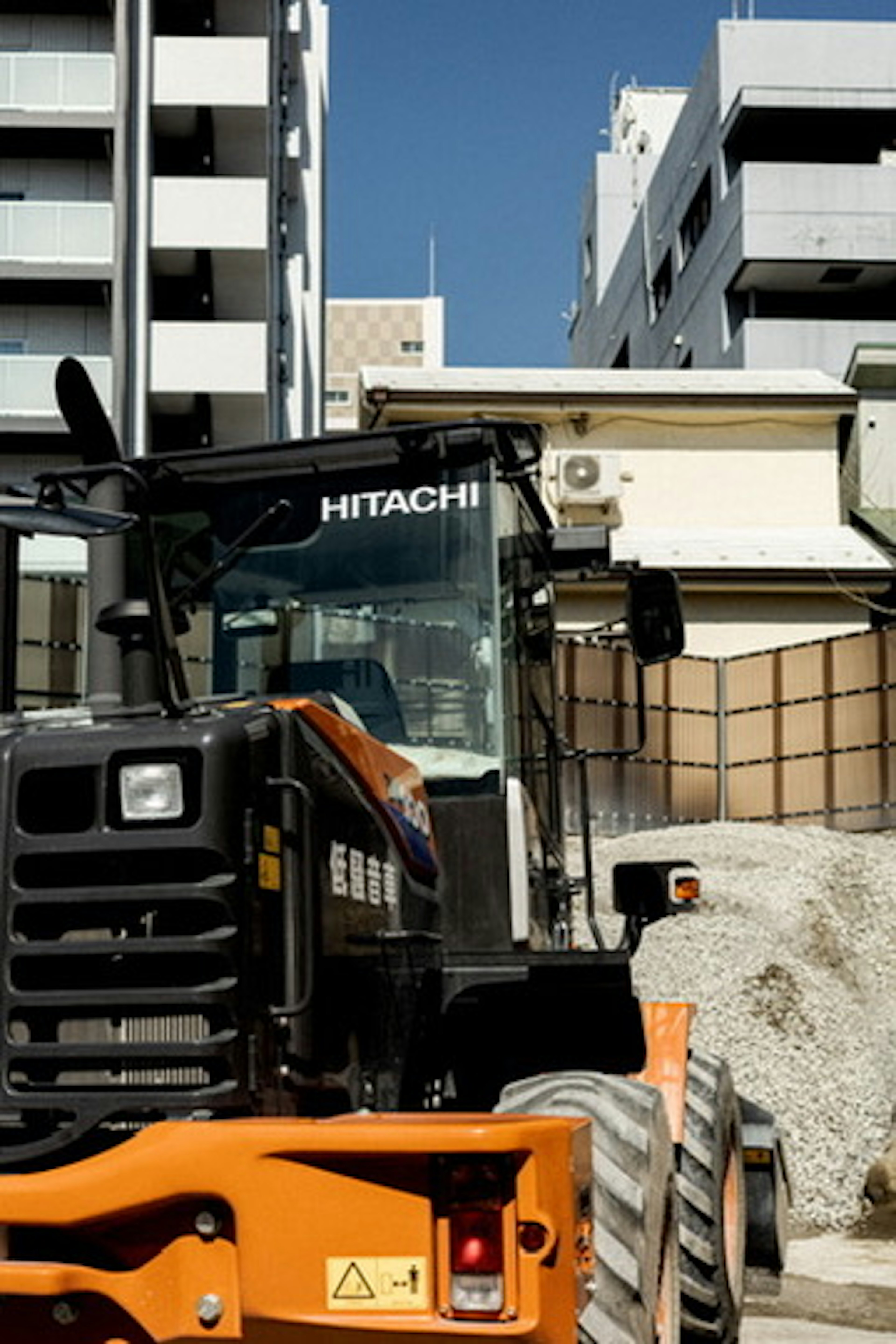 Sebuah mesin konstruksi Hitachi dengan trek dan bangunan di latar belakang