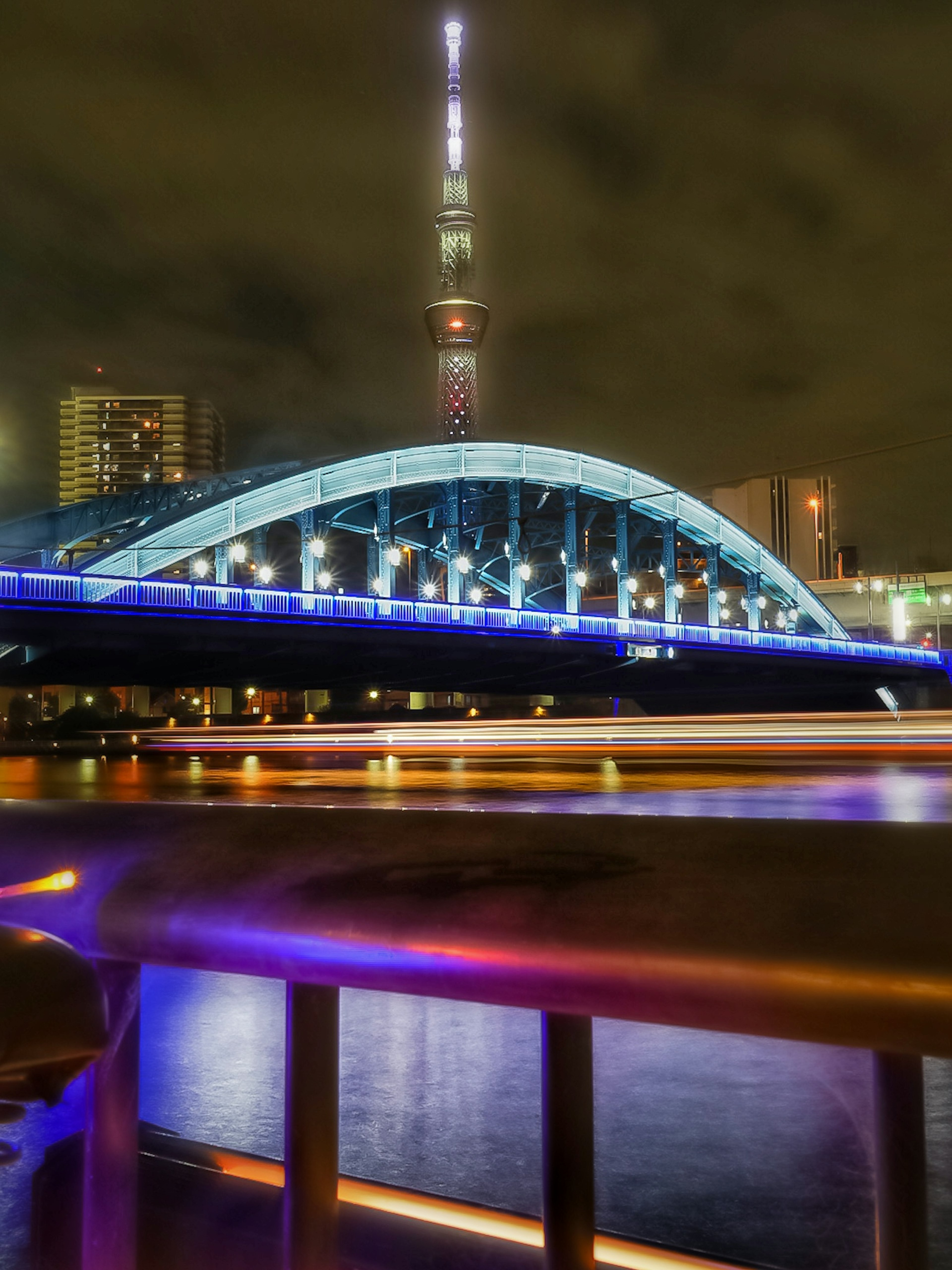 Vue nocturne d'un pont avec la Tokyo Skytree en arrière-plan éclairée en bleu et violet