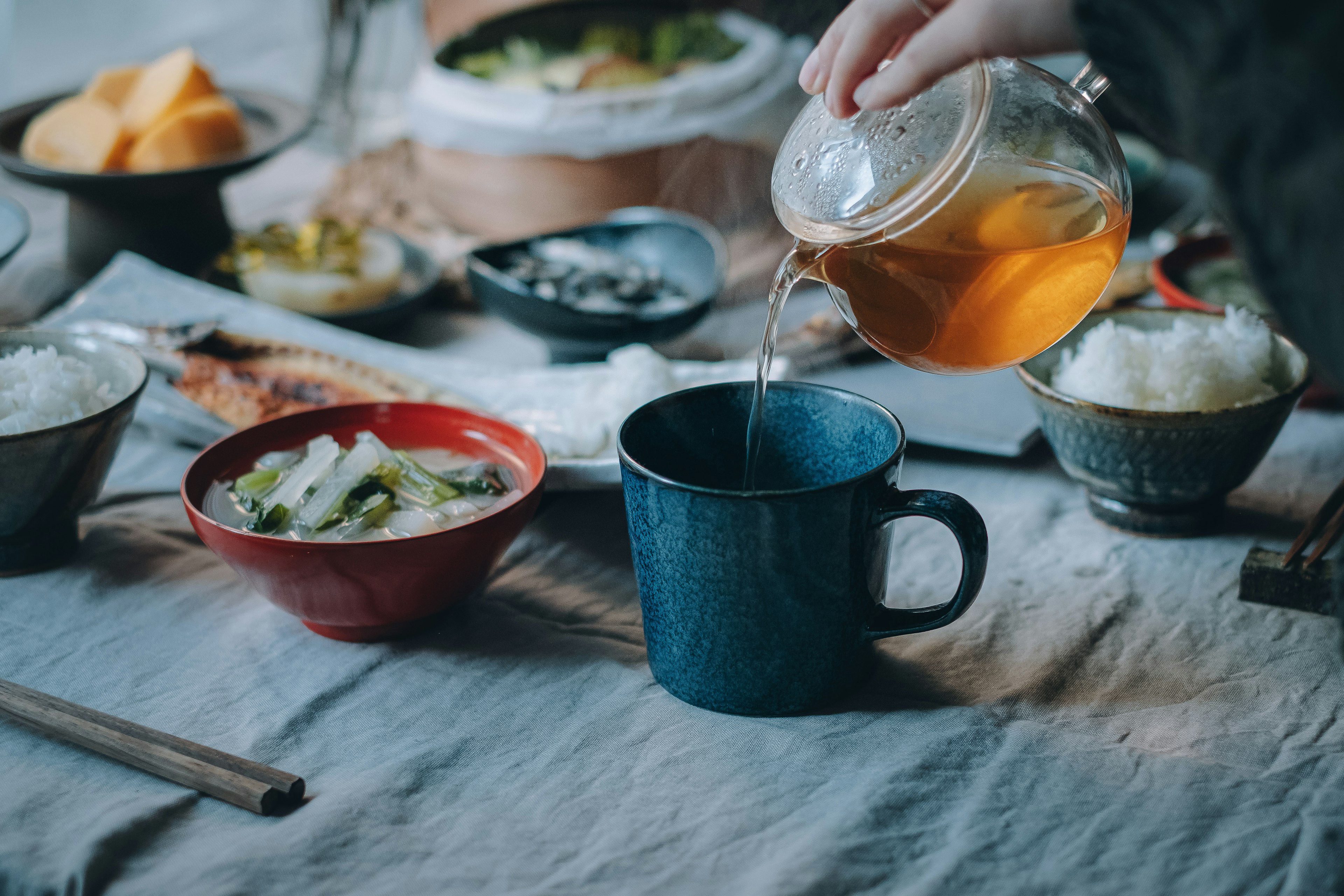 青いマグカップにお茶を注ぐ手と様々な料理が並んだテーブル