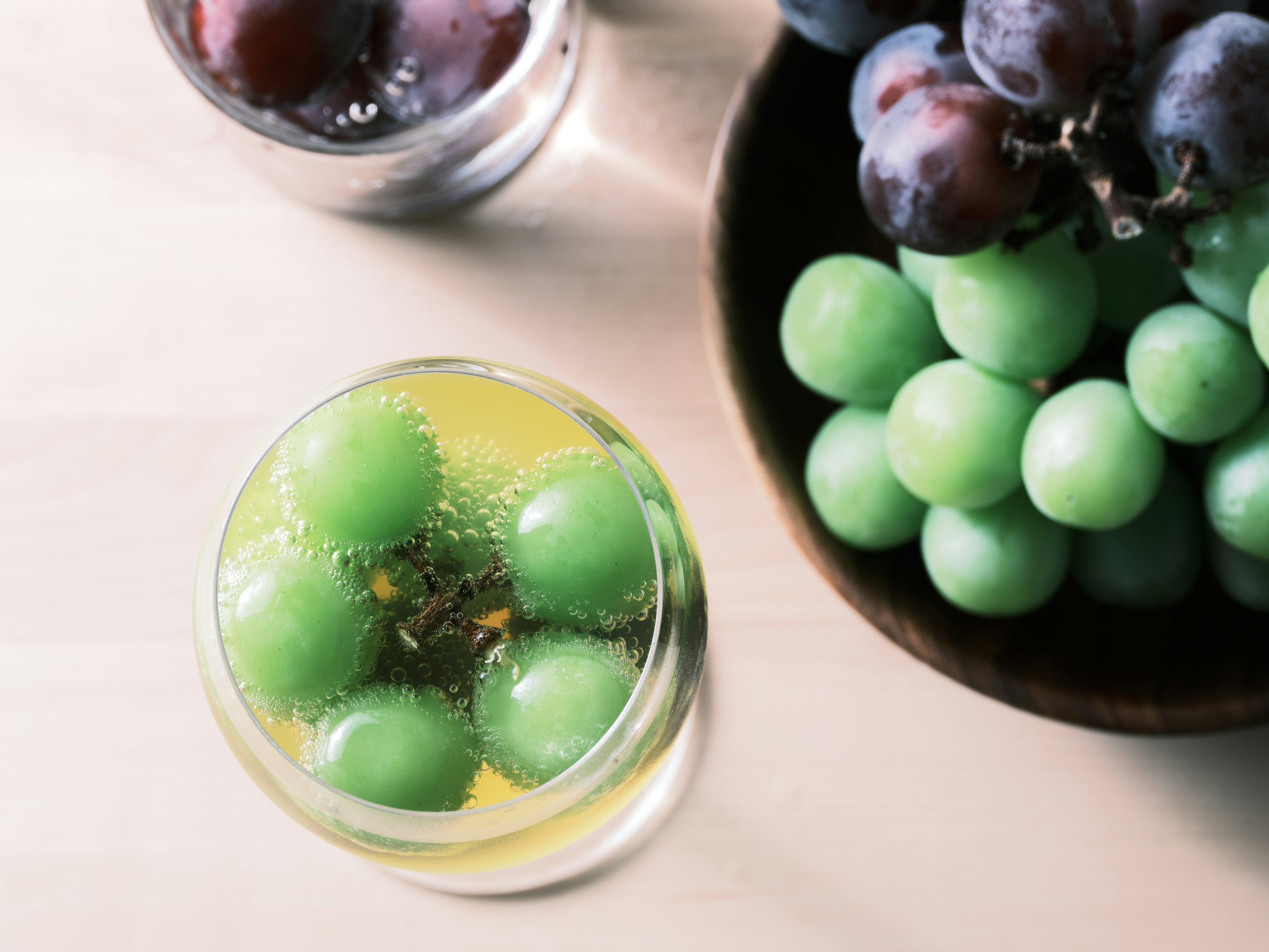 A glass of cocktail with green grapes and a bowl of grapes on a wooden table