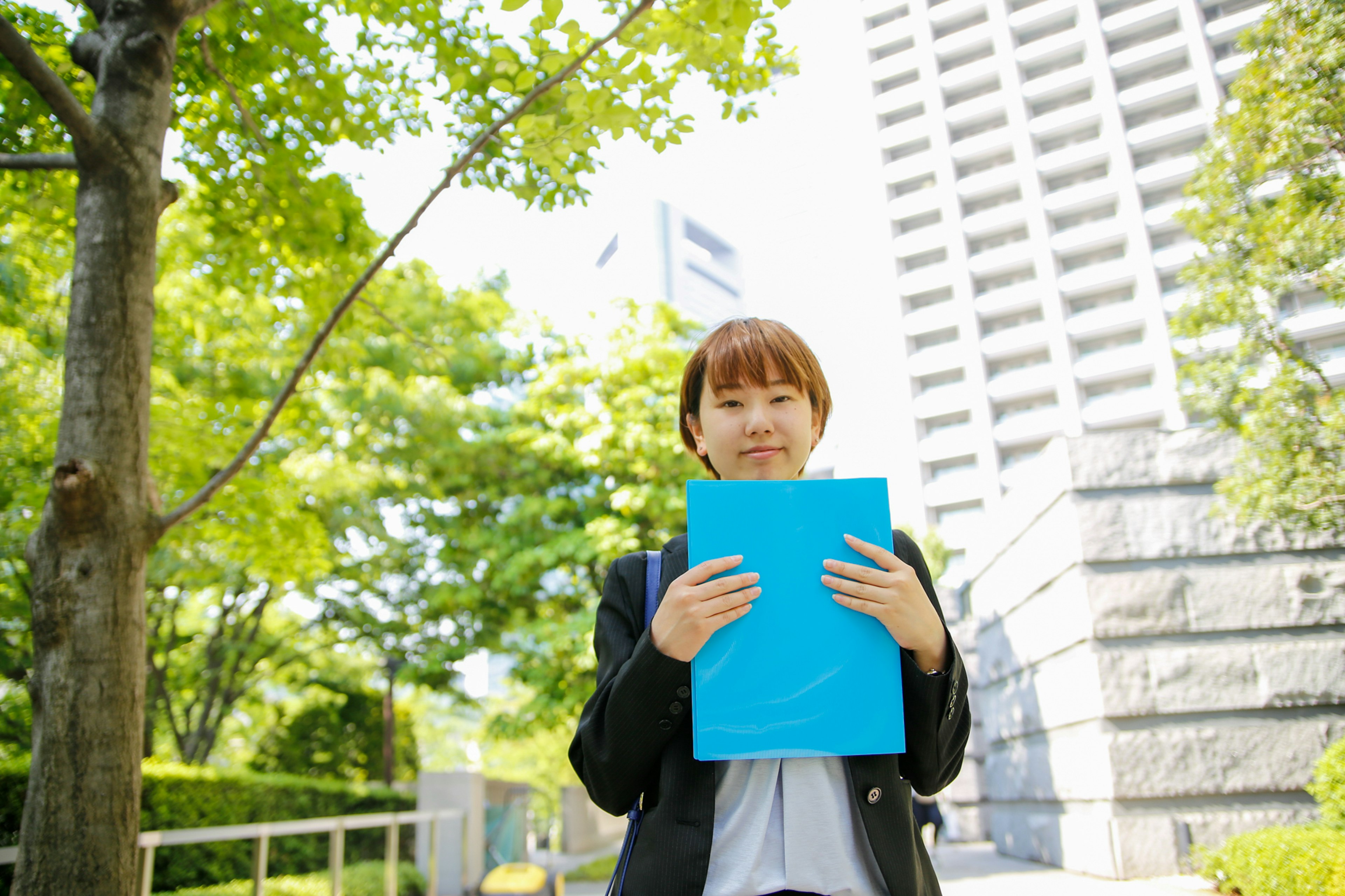 A woman in a business suit holding a blue folder standing in front of green trees and a high-rise building