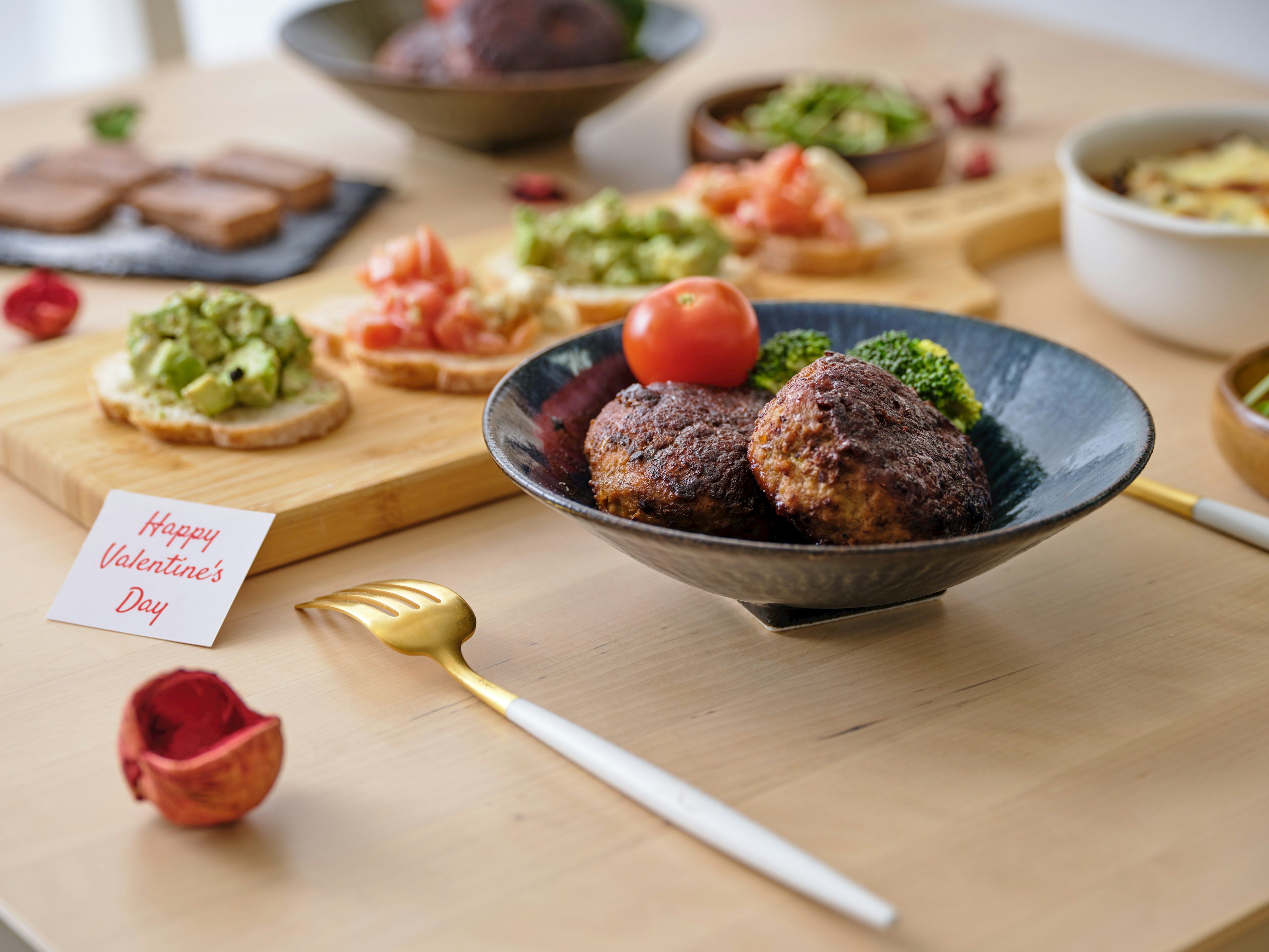 Presentación de comida temática de San Valentín con albóndigas y verduras frescas en un tazón negro rodeadas de tostadas y ensalada