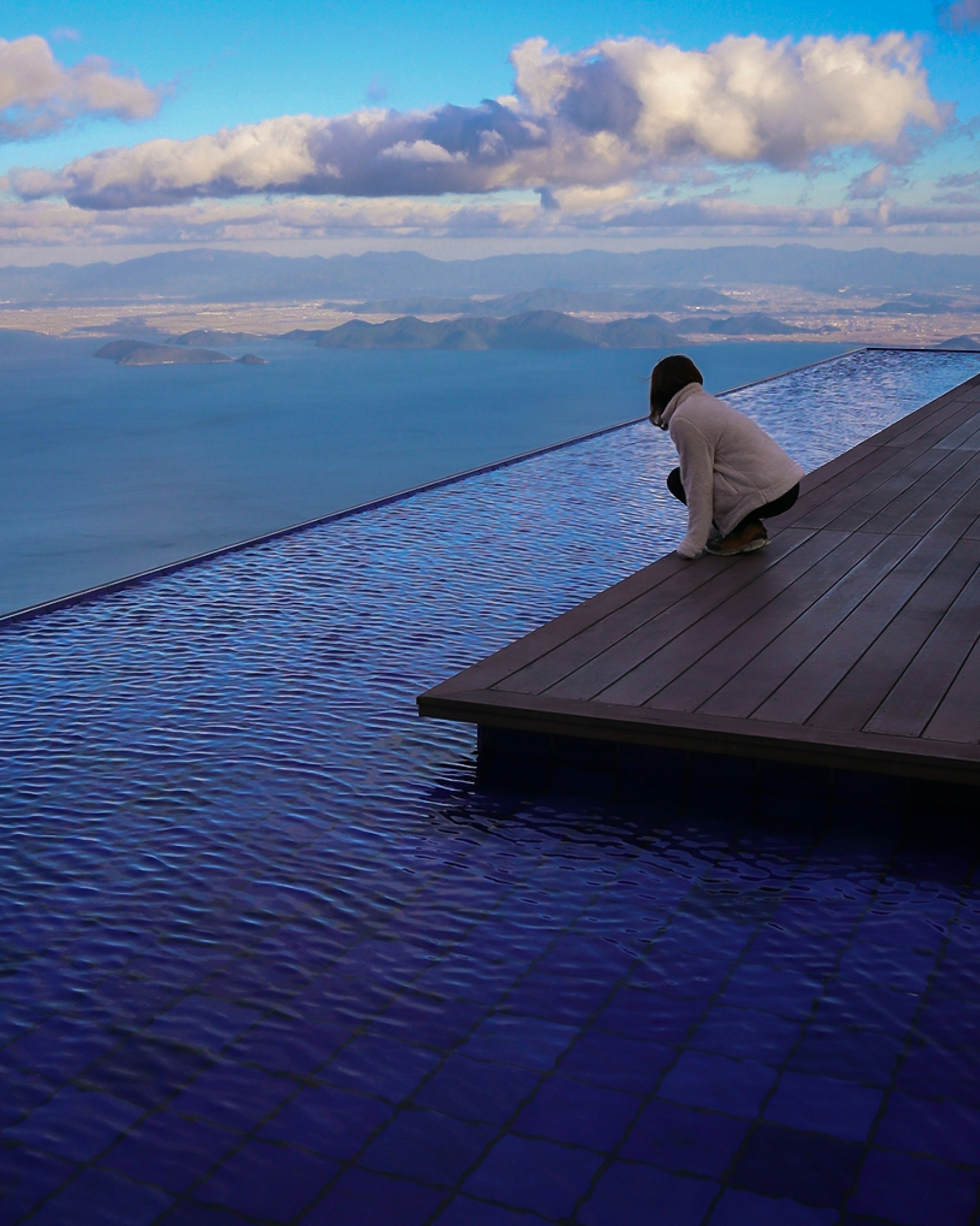 Persona seduta sul bordo di una piscina a sfioro che guarda il cielo blu e l'acqua