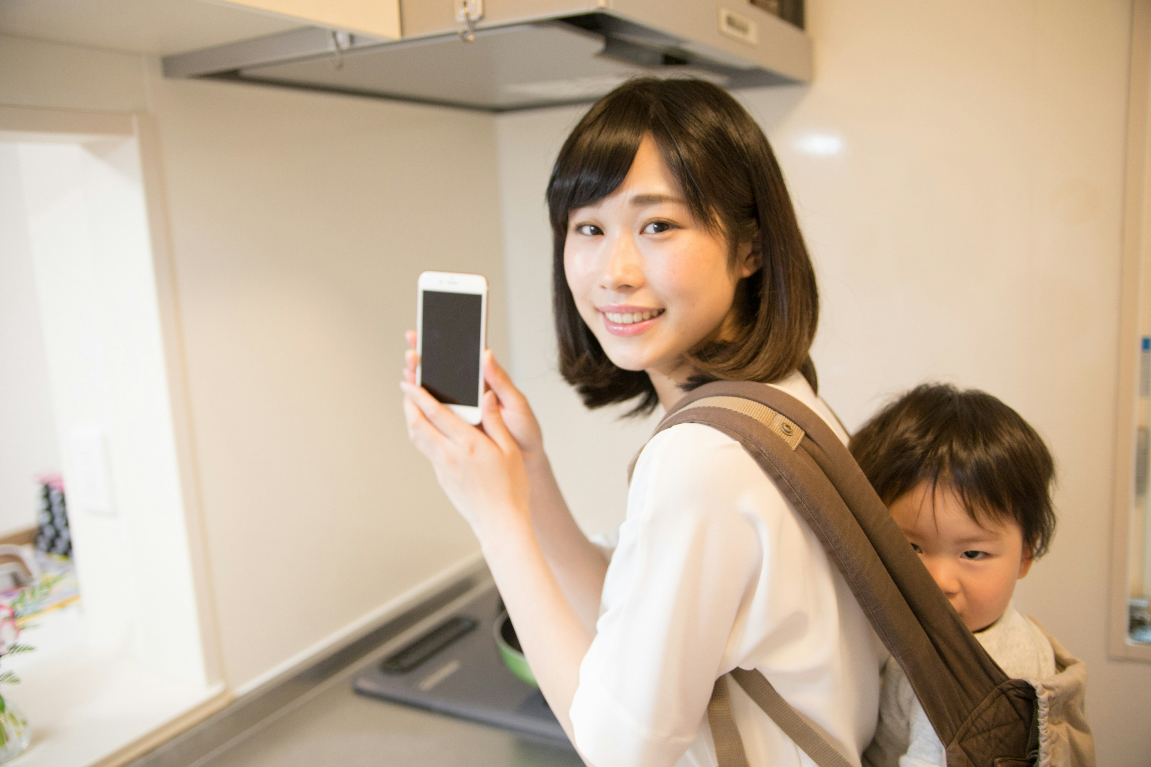 Woman holding a smartphone in a kitchen with a child on her back