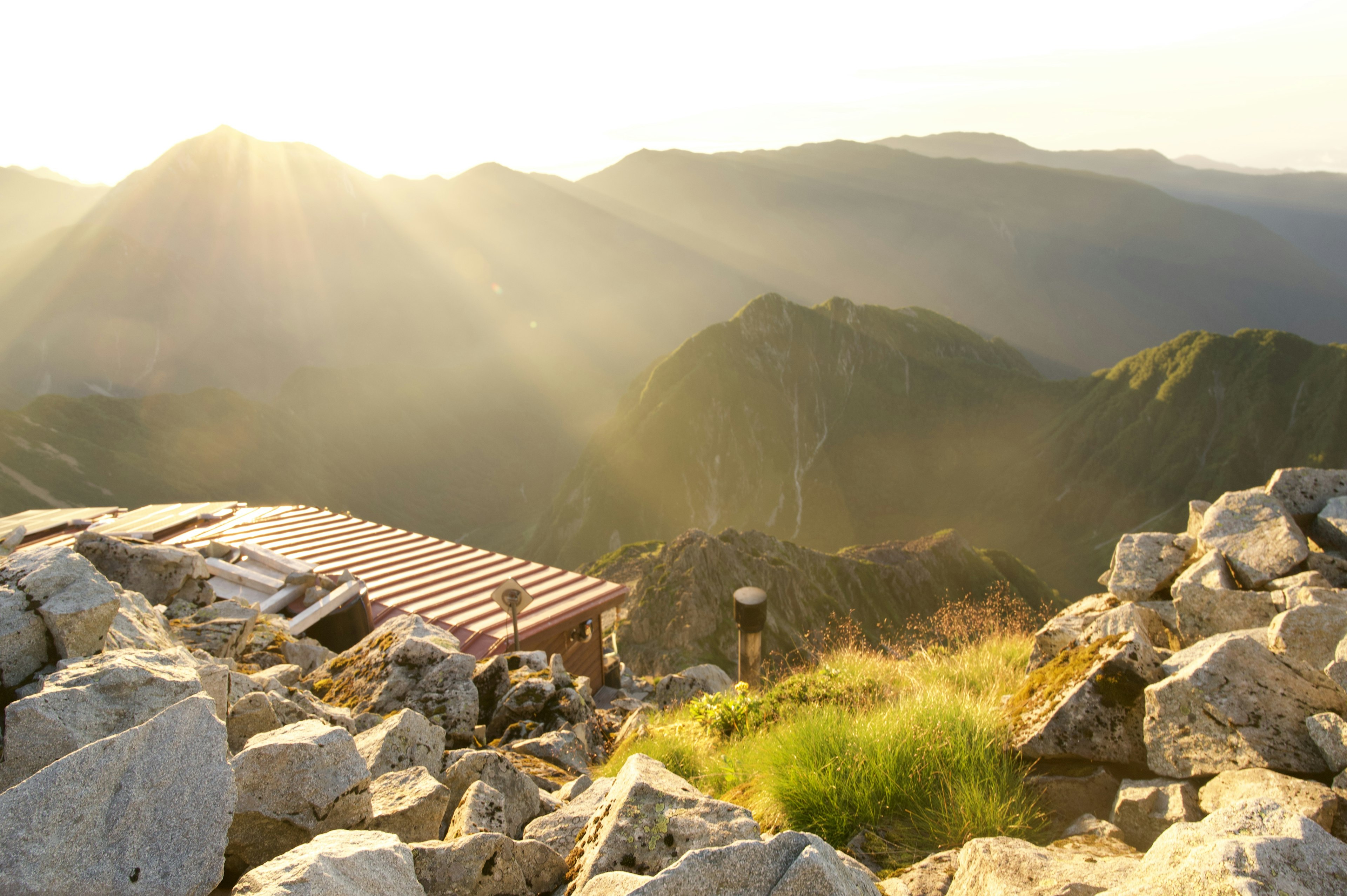 Panoramablick von einem Berggipfel mit einer Hütte umgeben von Felsen und Gras beleuchtet von Sonnenuntergang