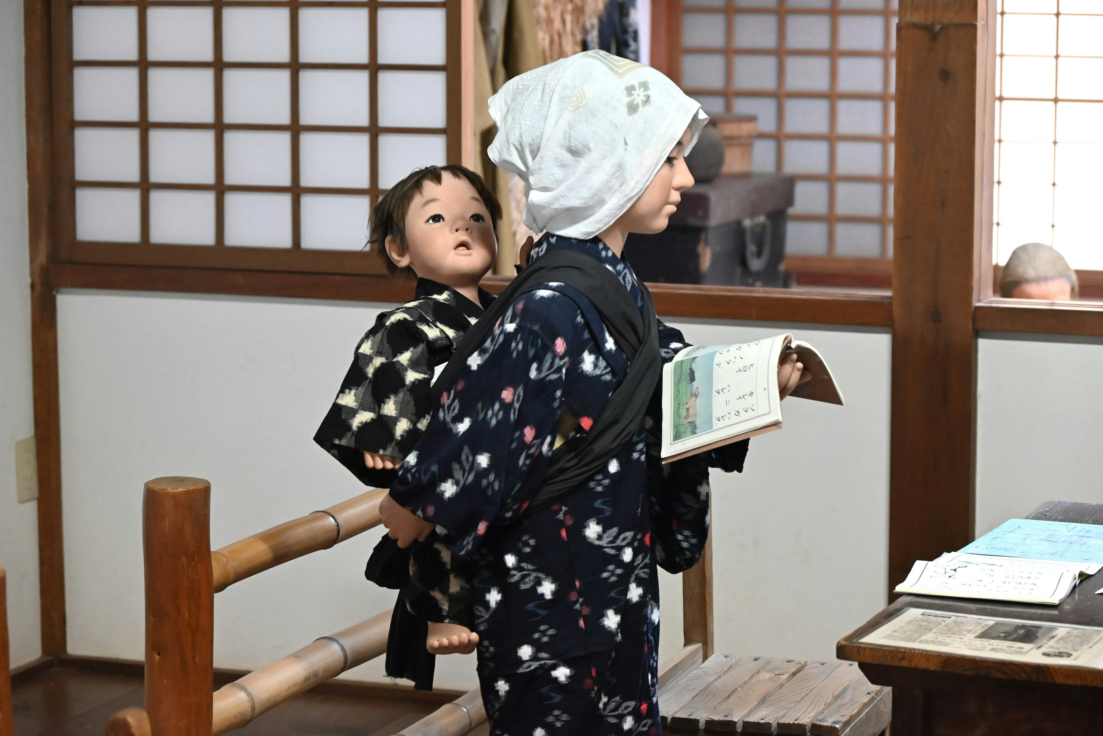 Ein Junge im Kimono, der eine Puppe auf dem Rücken trägt, hält ein Buch in einem traditionellen japanischen Raum