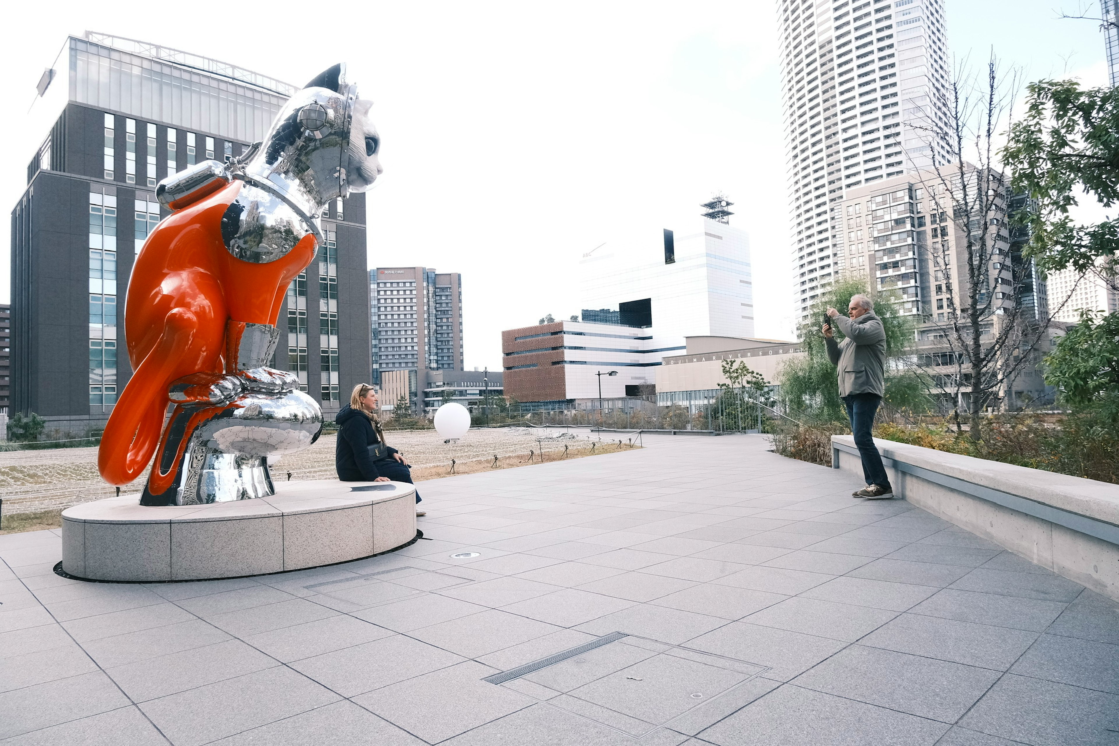 A silver and orange dog sculpture in a park with surrounding buildings