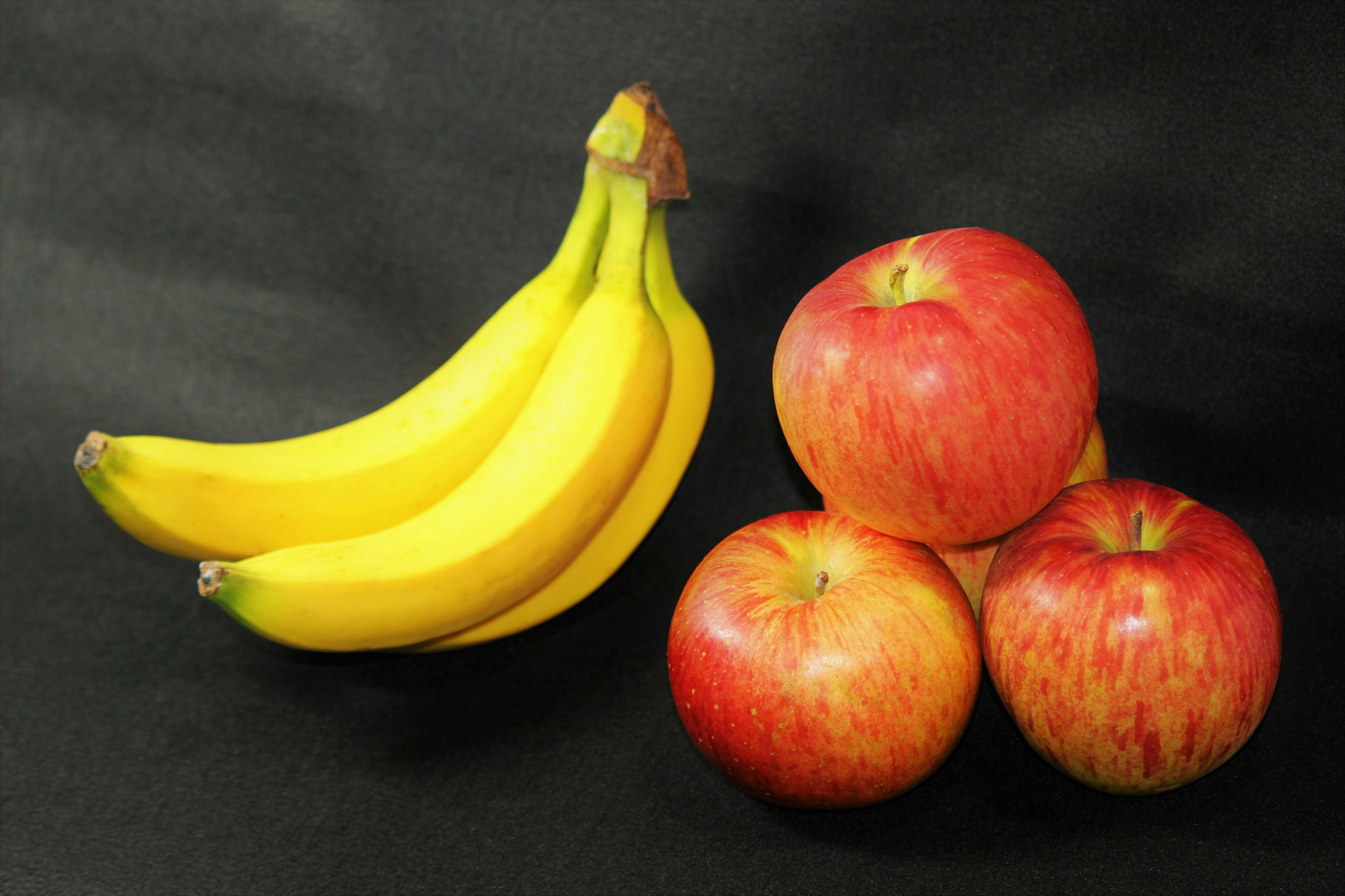 A bunch of yellow bananas next to a group of red apples on a black background