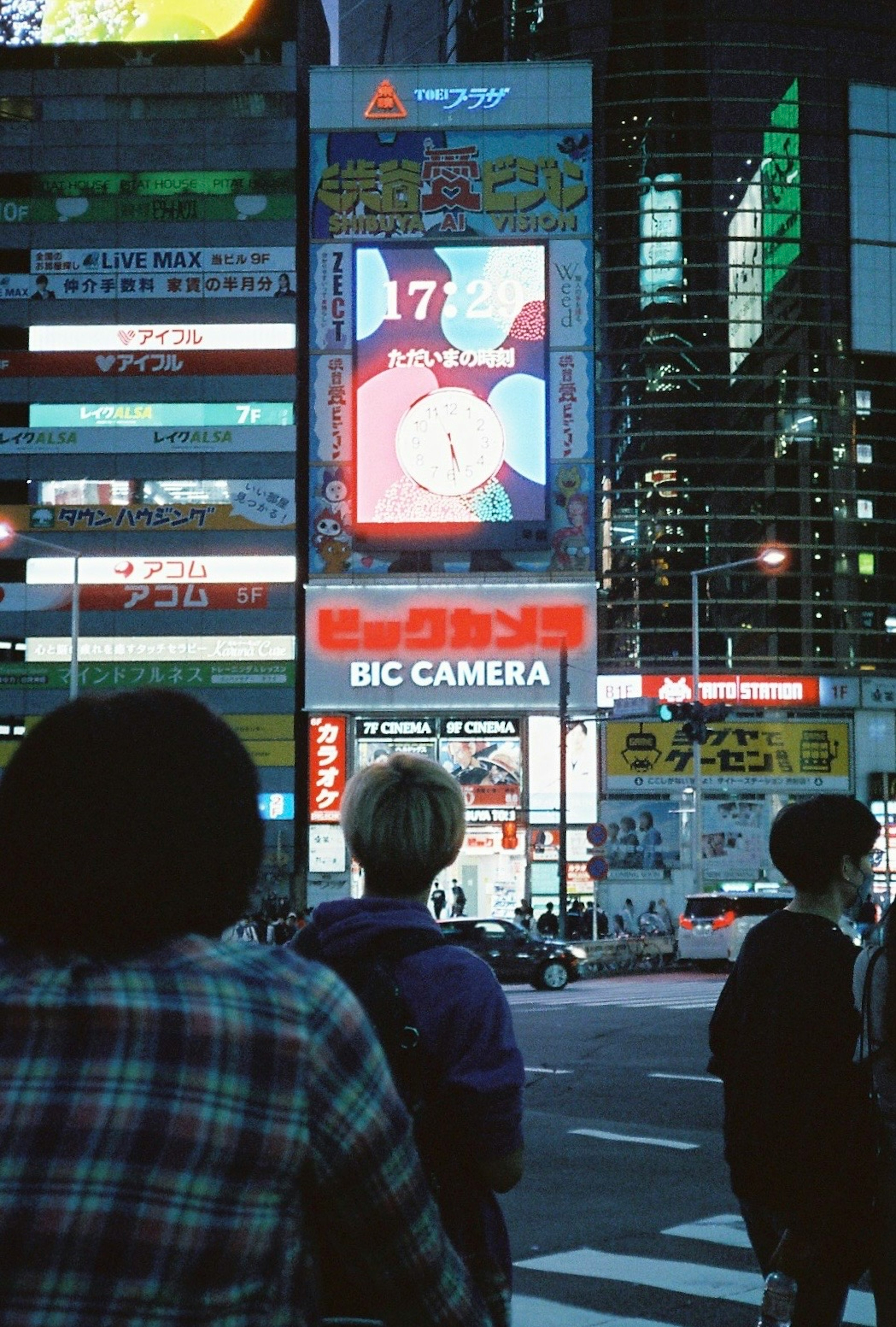 Escena nocturna en un área bulliciosa con un letrero de Bic Camera la gente mirando un anuncio de anime