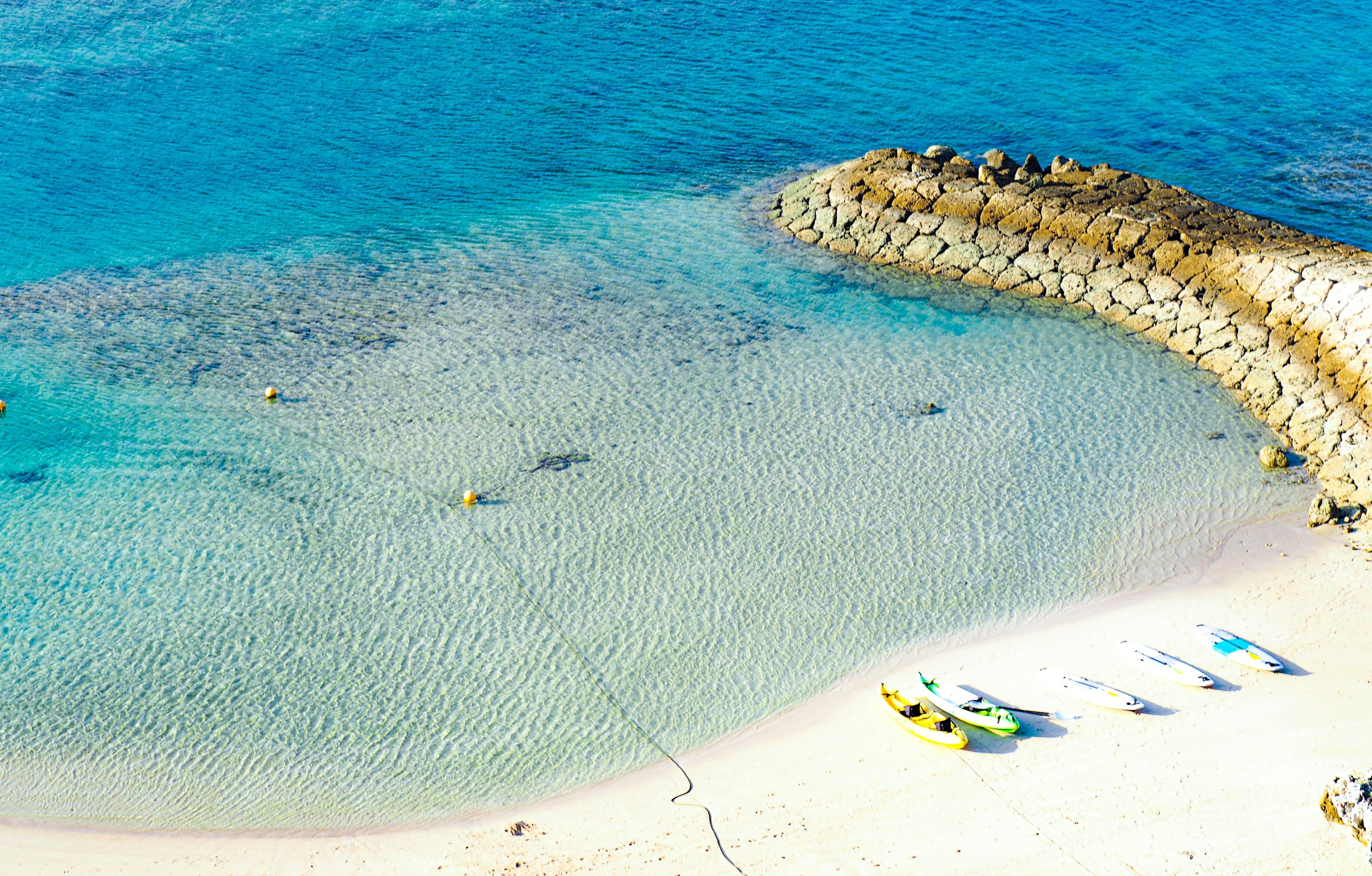 Aerial view of a beach with clear blue water and sandy shore featuring a rocky jetty and colorful boats