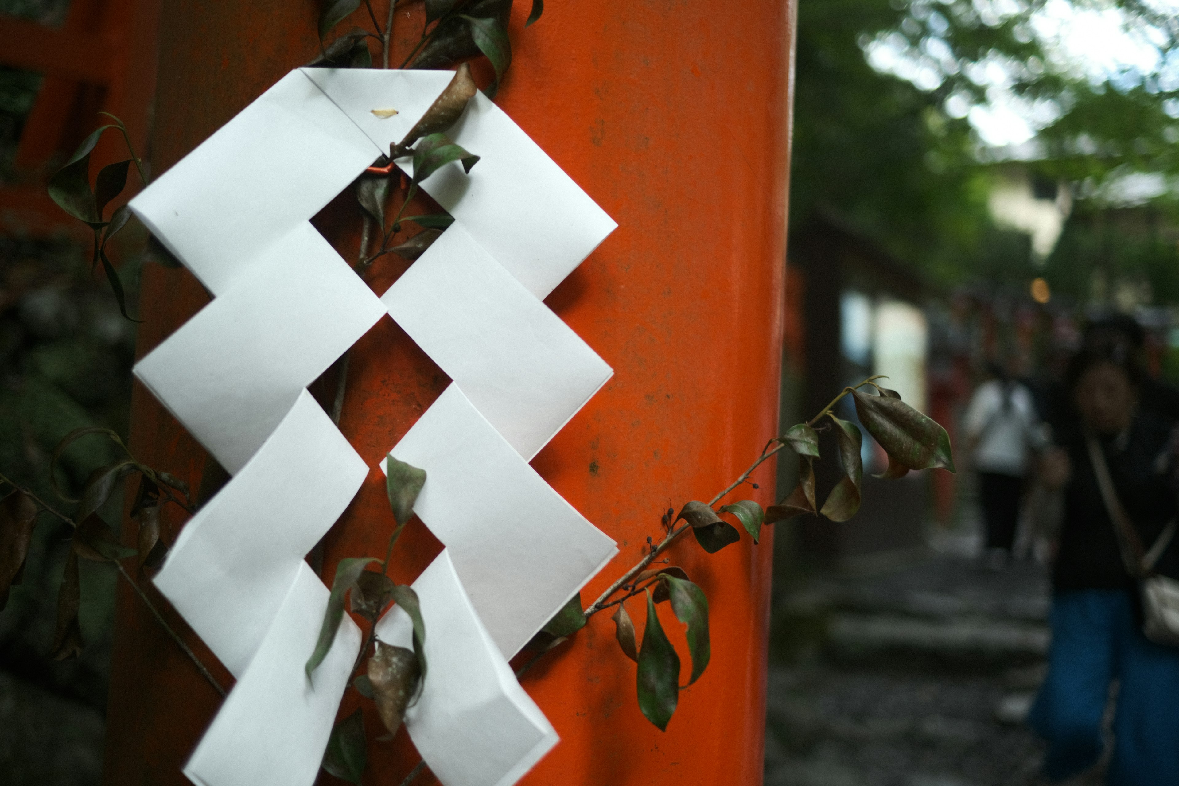 Decorazione in carta bianca su un torii arancione con persone che camminano sullo sfondo