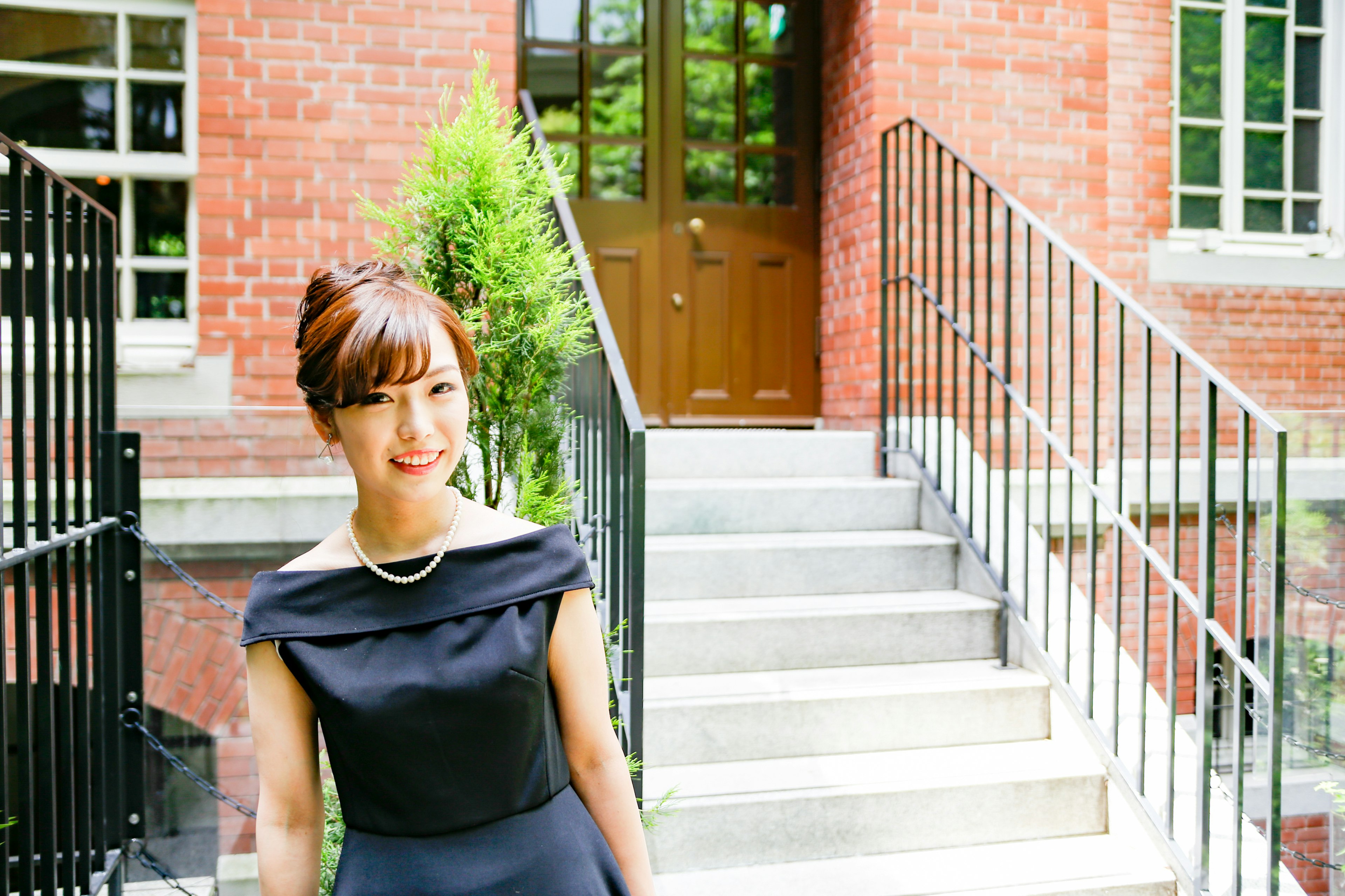 A woman in a black dress standing in front of stairs with a brick building and greenery in the background