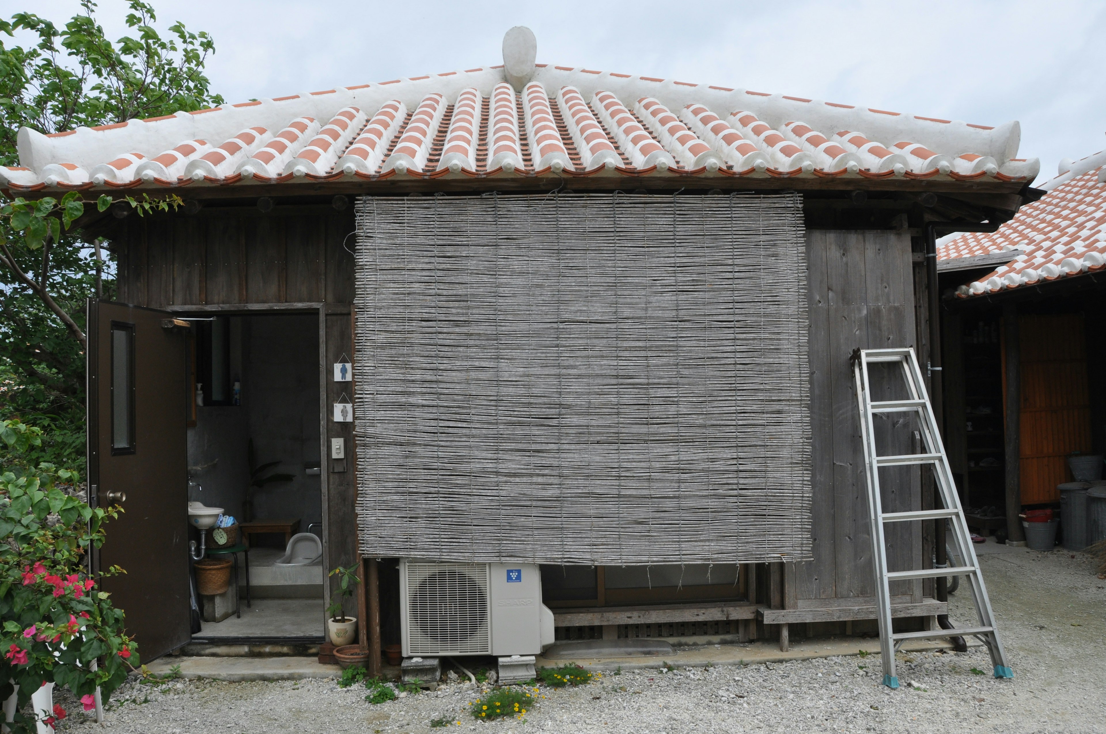 Ancienne maison en bois avec toit en tuiles rouges, fenêtre couverte de tissu gris, échelle appuyée contre le mur