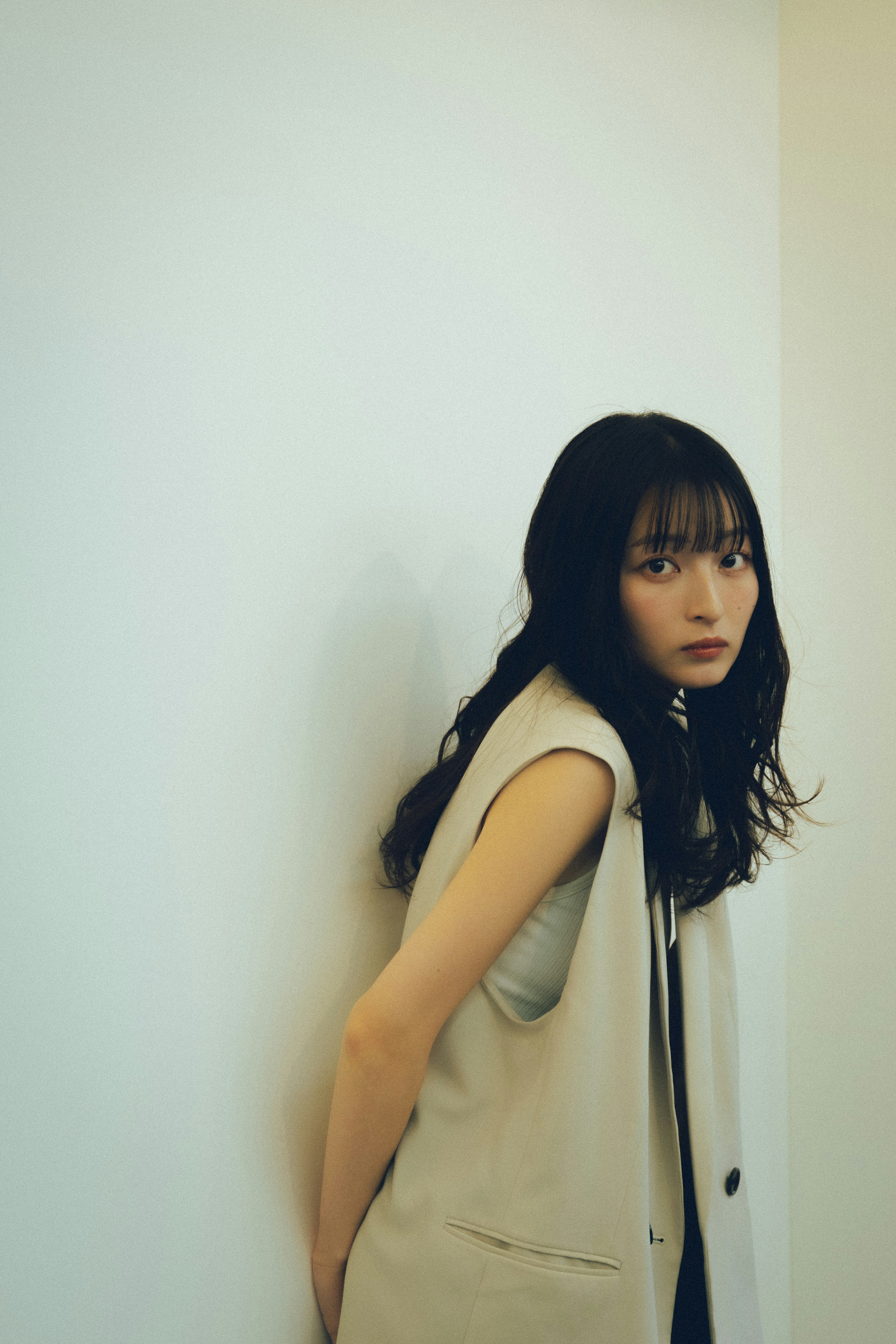 Portrait of a woman leaning against a wall with long black hair wearing a beige vest