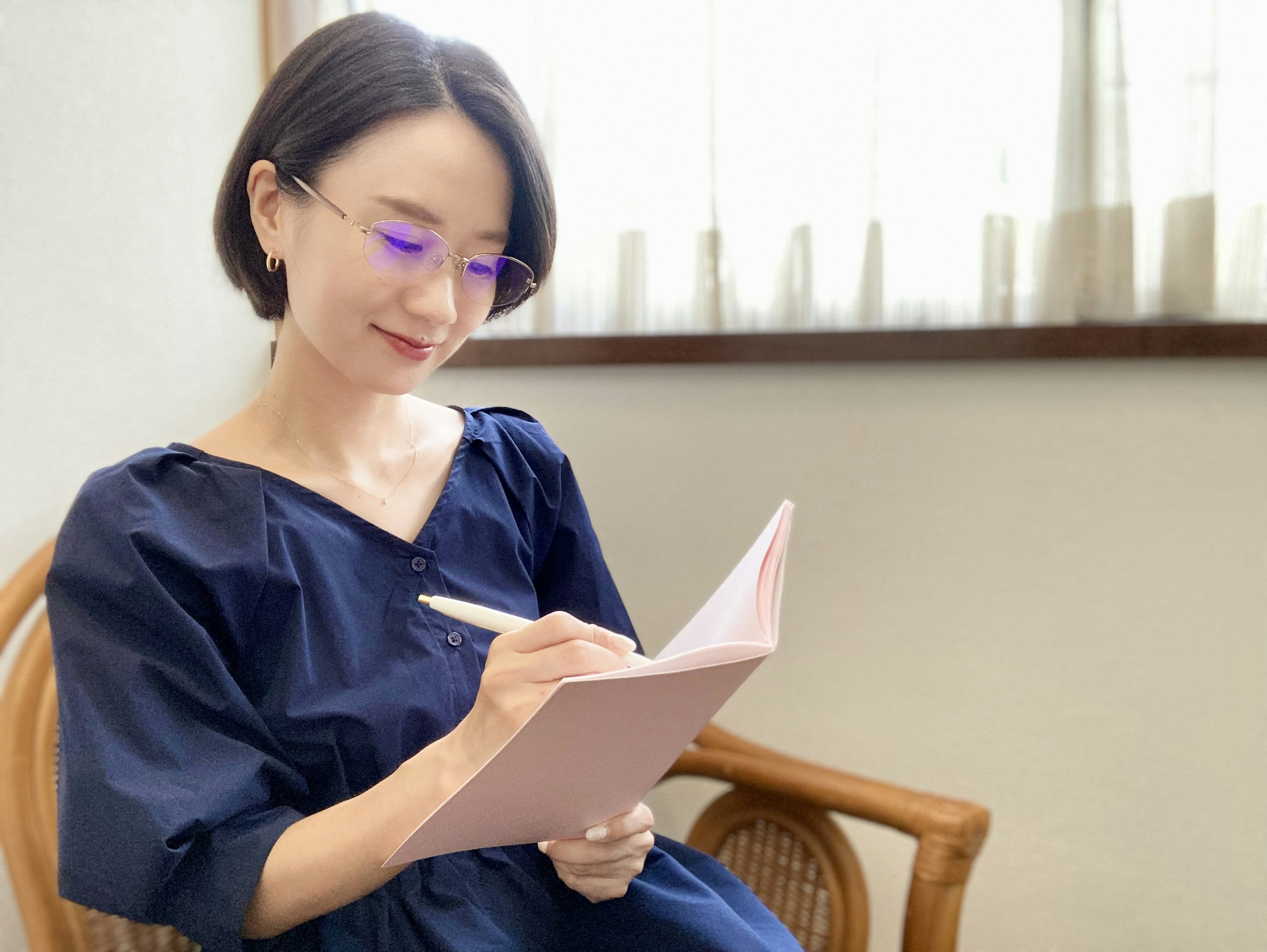 Mujer escribiendo en un cuaderno junto a la ventana con un vestido azul y gafas moradas en un ambiente relajado