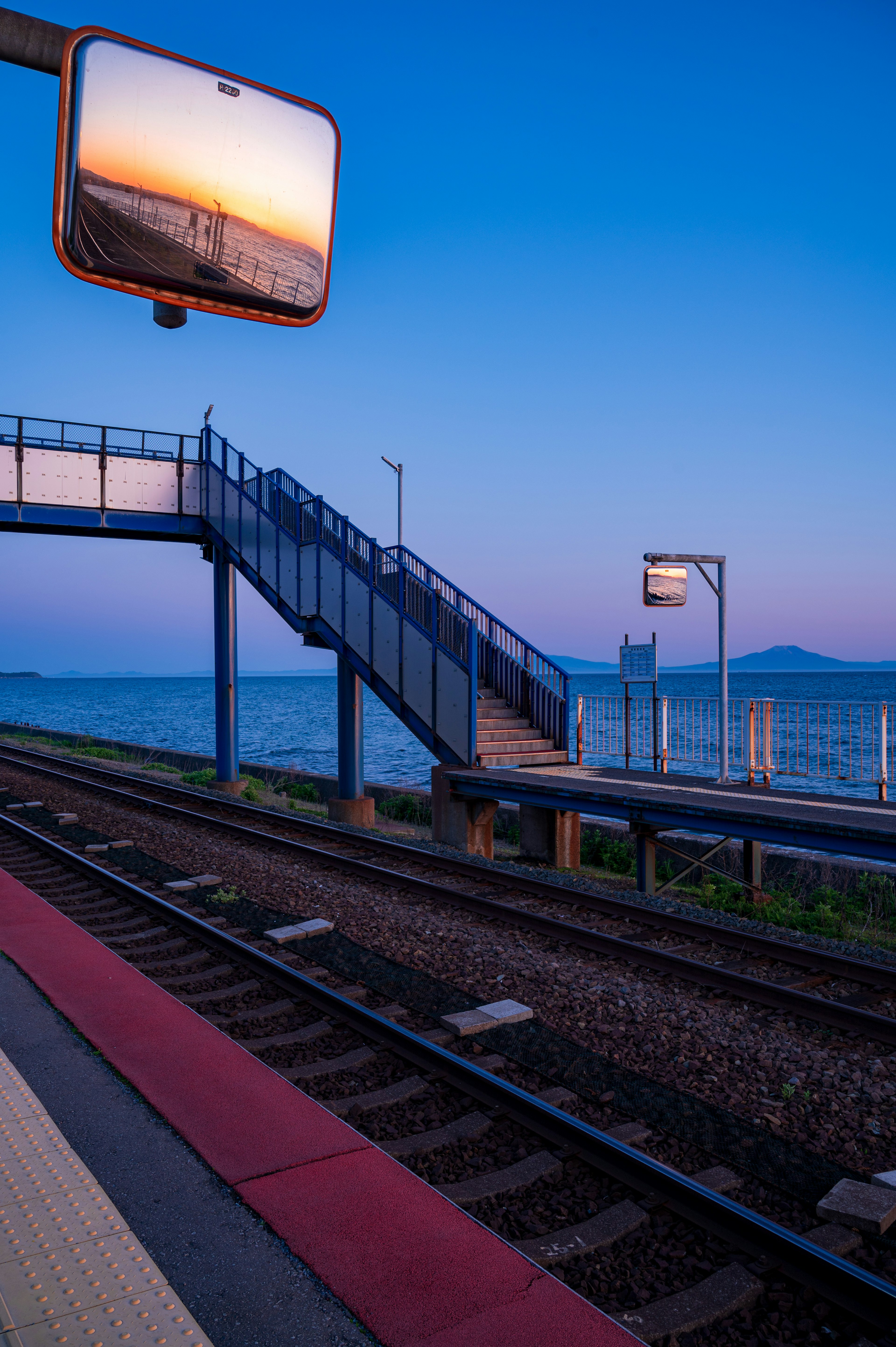 Plataforma de estación de tren con escaleras que dan al mar reflejando el cielo del atardecer en un espejo