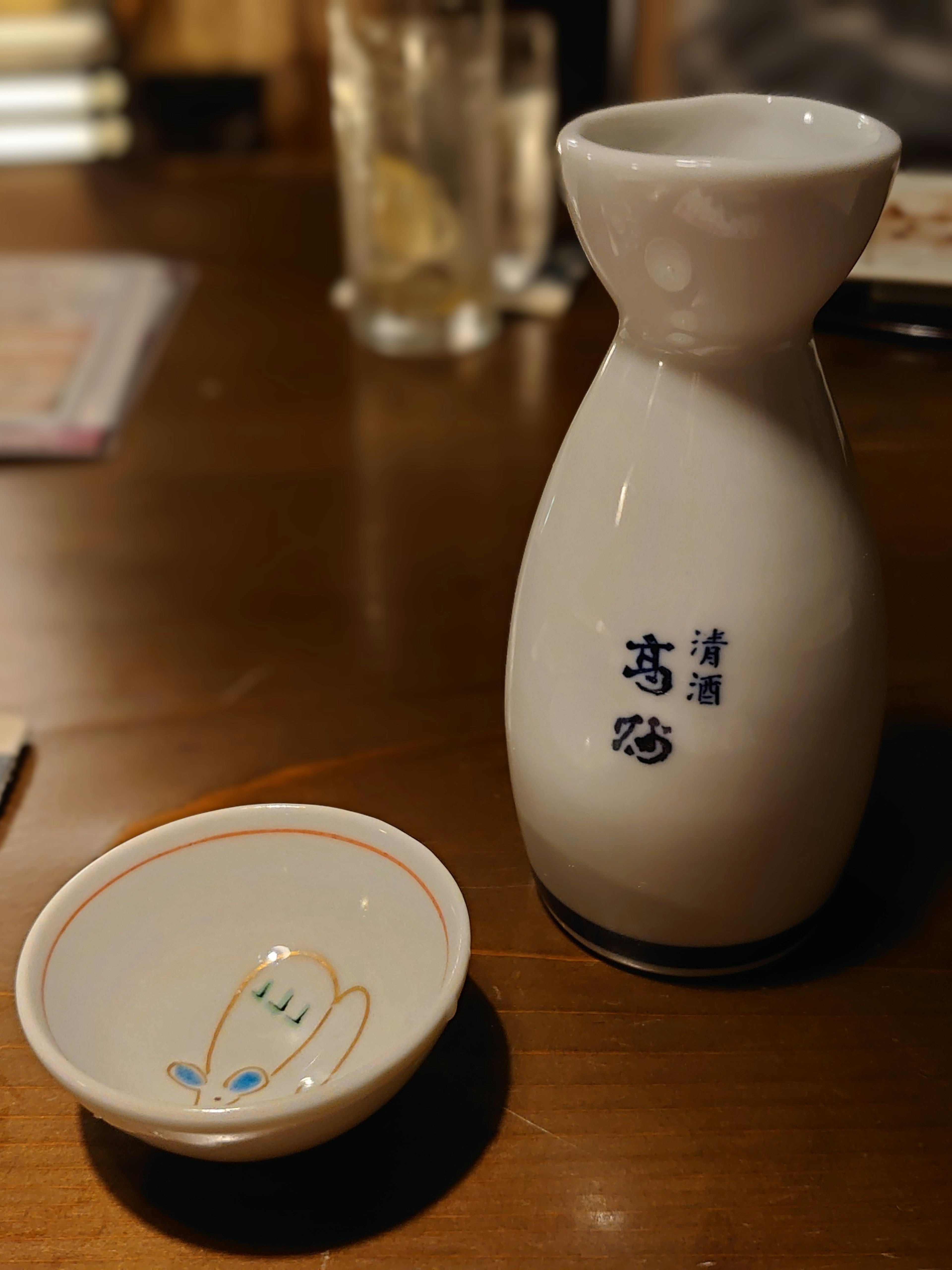 A white sake bottle with blue lettering and a small dish on a wooden table