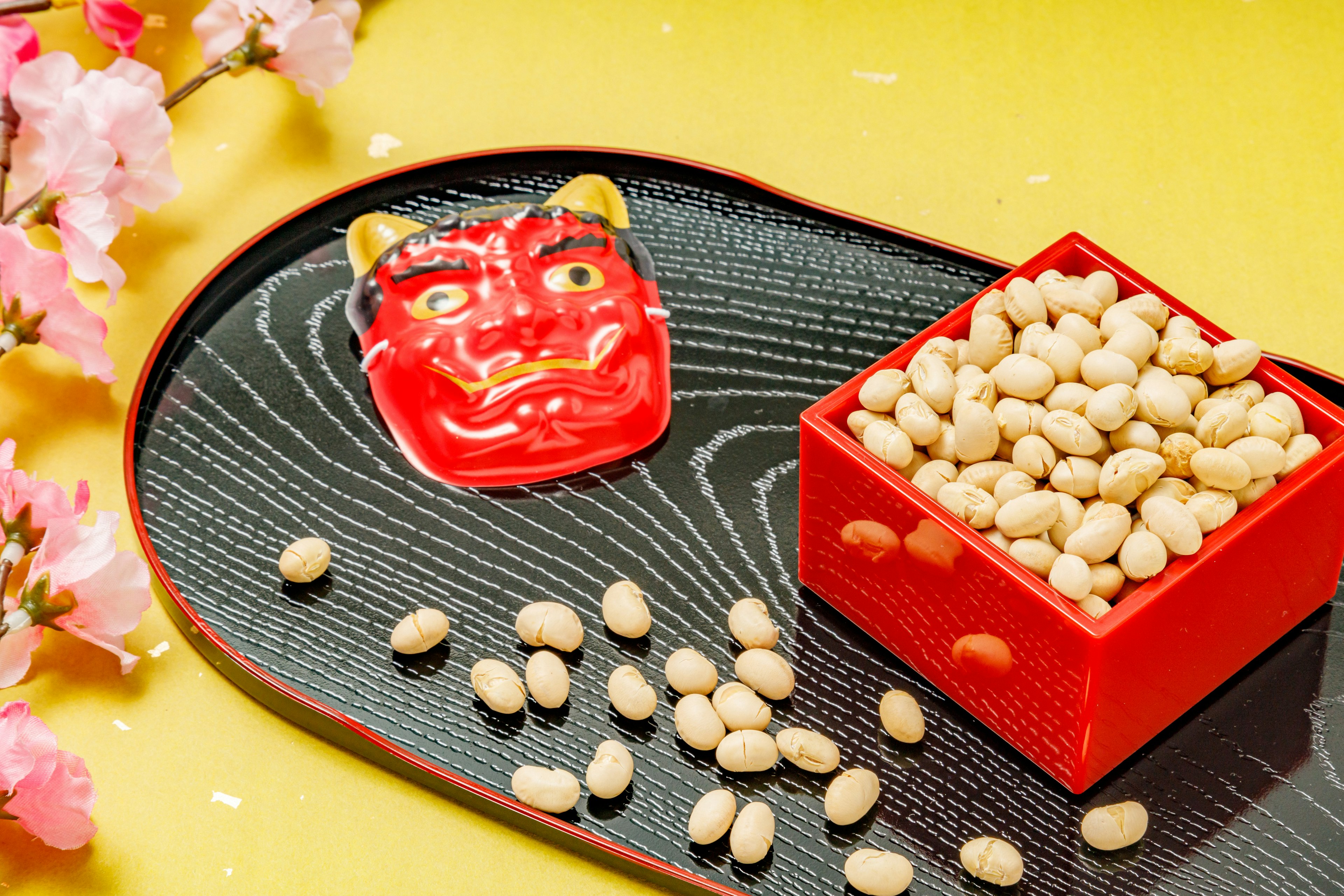 Máscara de demonio roja con una caja de frijoles sobre un plato de madera contra un fondo amarillo