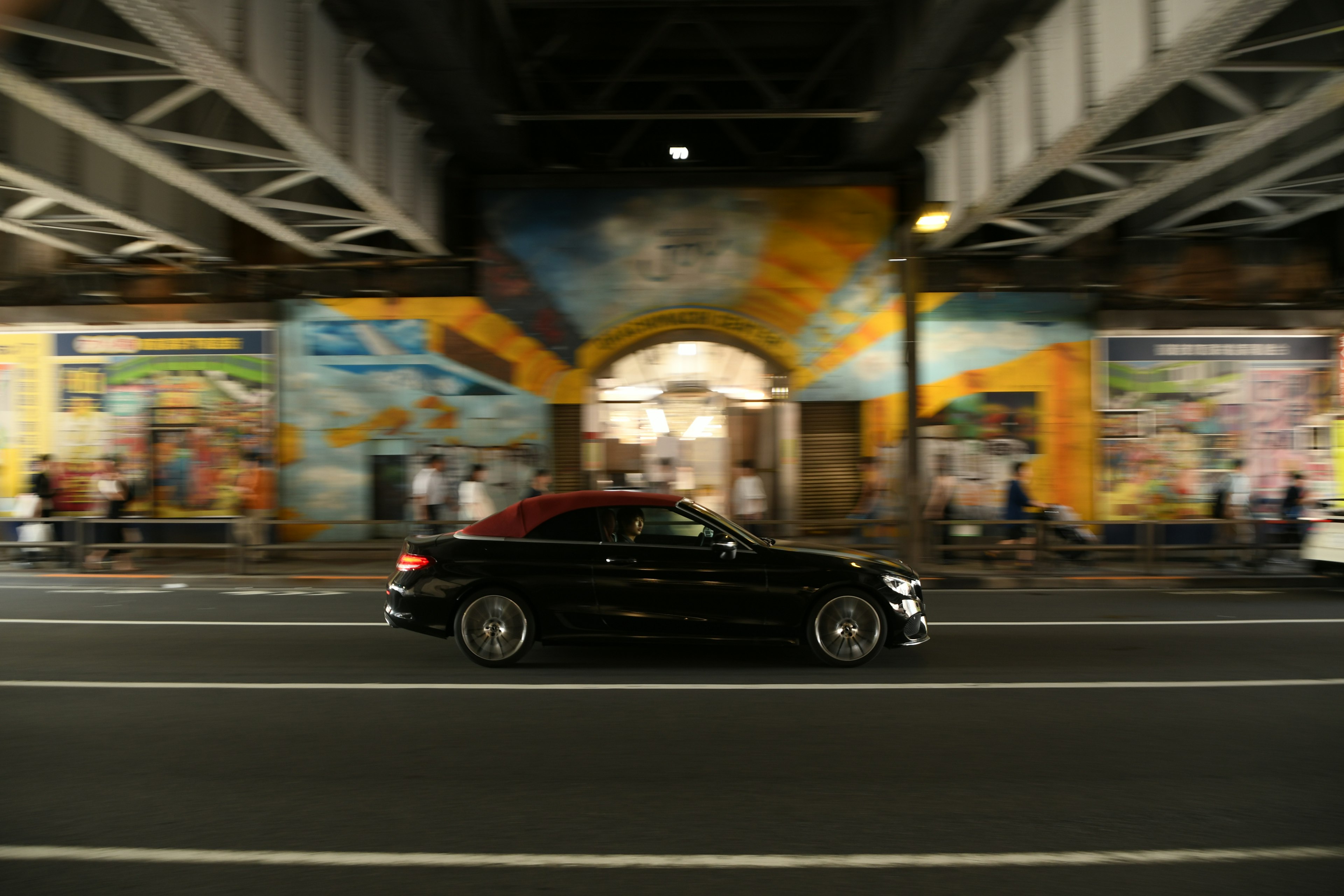 A black car driving past vibrant murals under a bridge