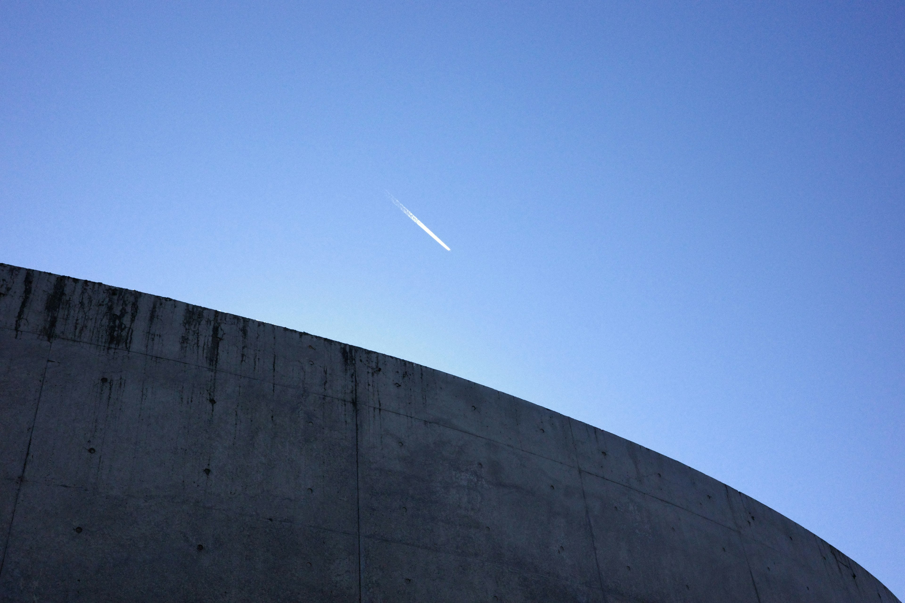 White contrail in blue sky above curved concrete structure