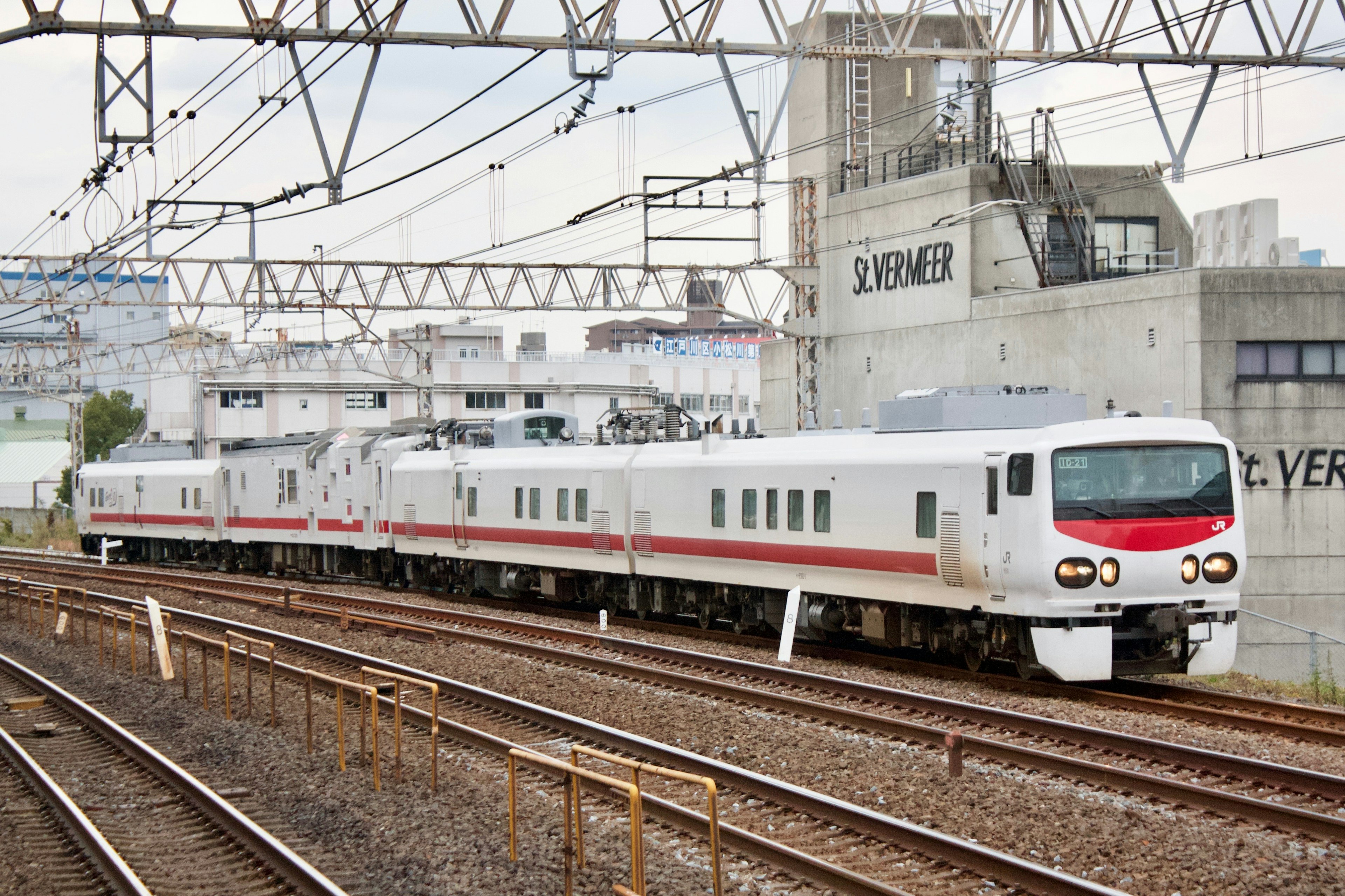 Un tren blanco que circula por las vías con edificios y líneas aéreas al fondo