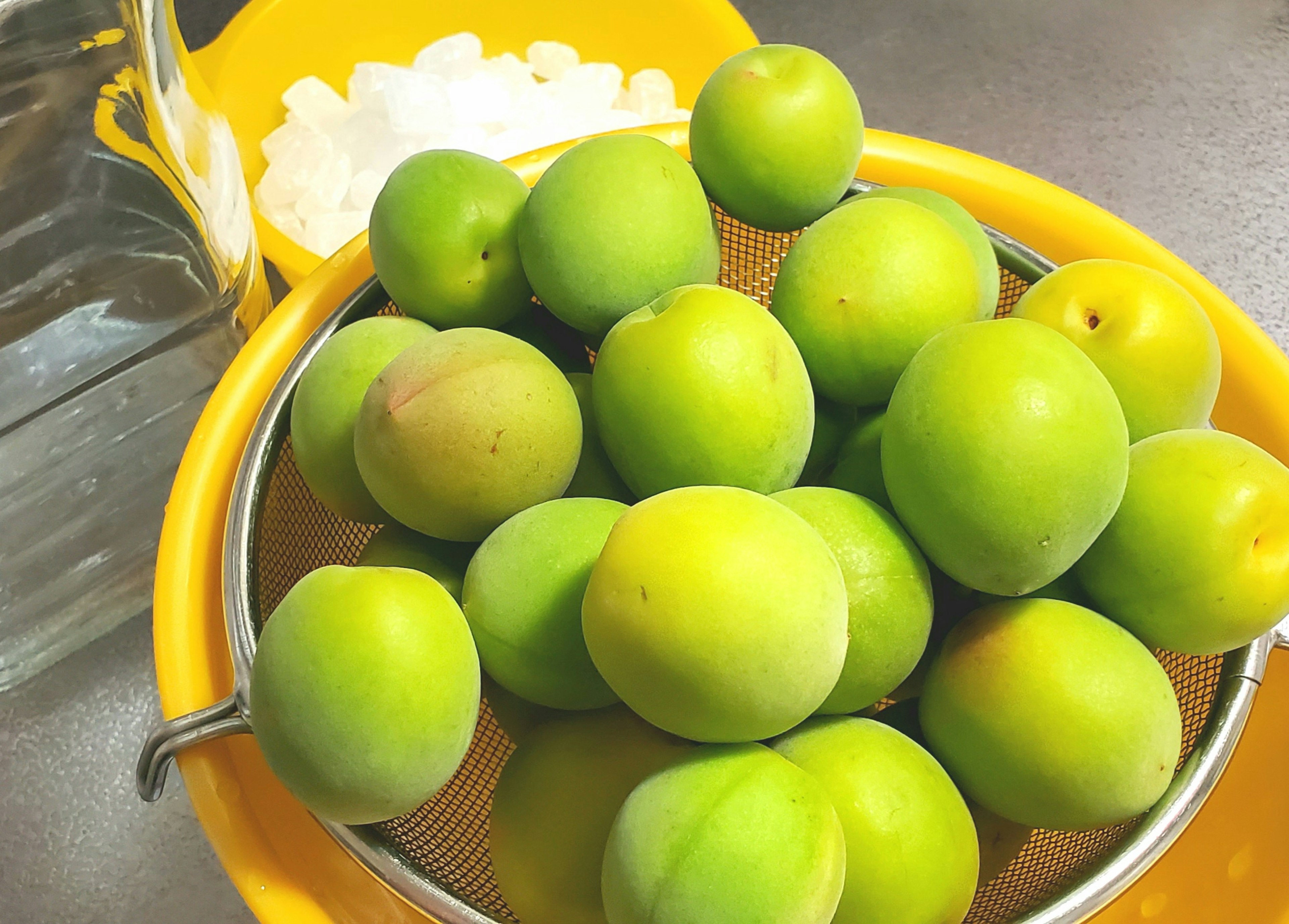 Sebuah mangkuk berisi plum hijau dalam saringan dengan toples transparan di latar belakang