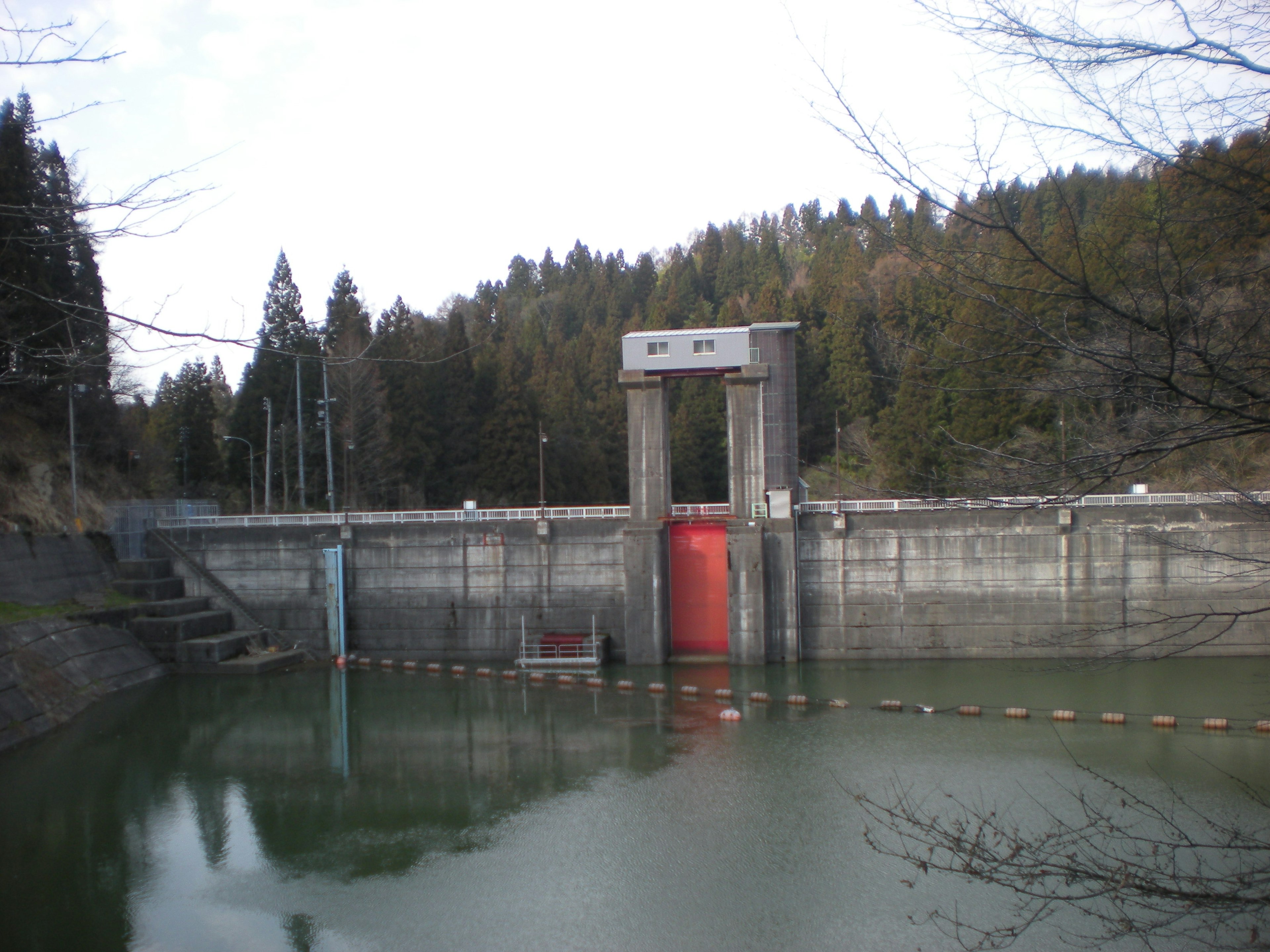 Dam with a red gate and surrounding forest reflected in calm water