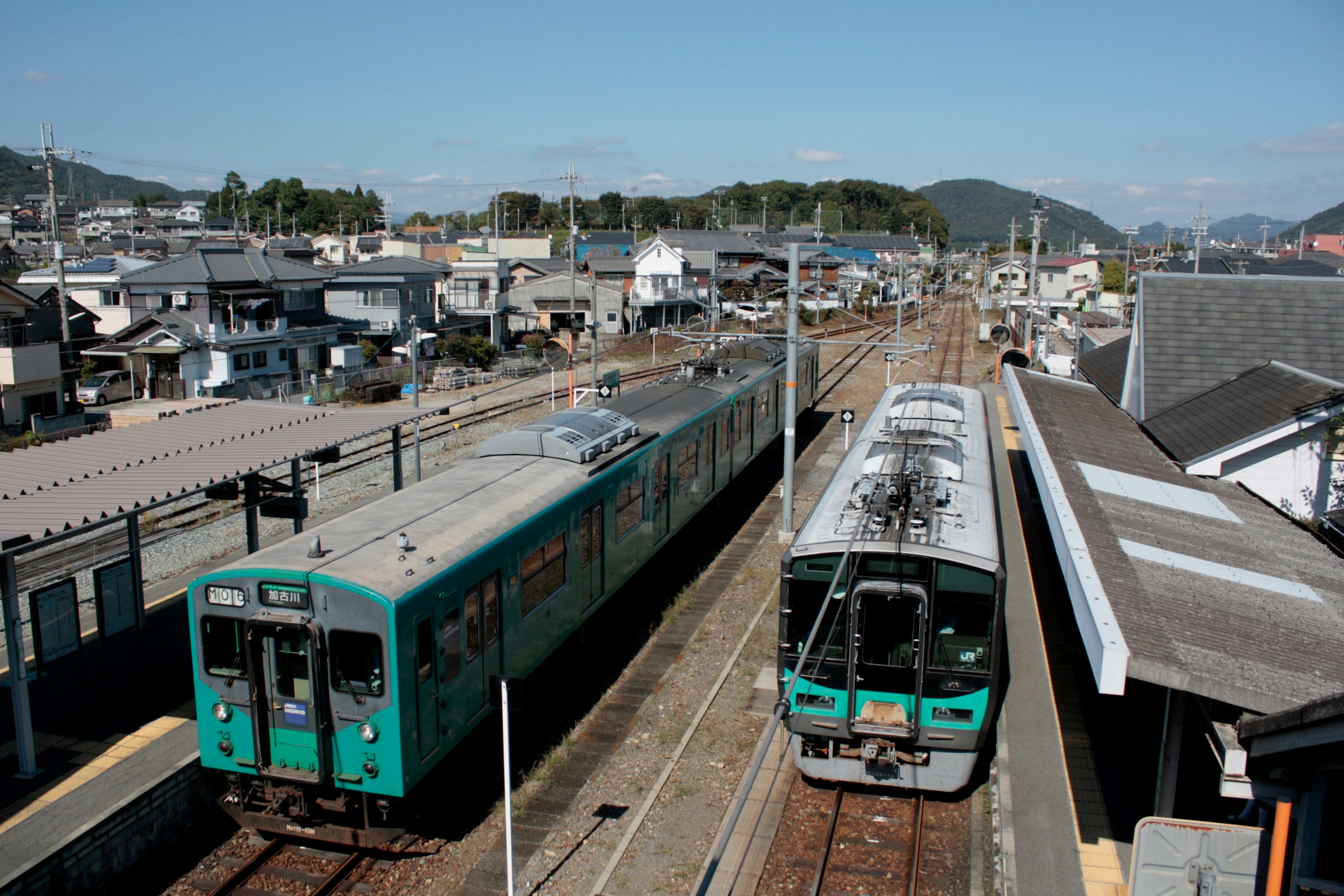 Sebuah kereta biru dan kereta hijau di stasiun dengan bangunan dan gunung di latar belakang