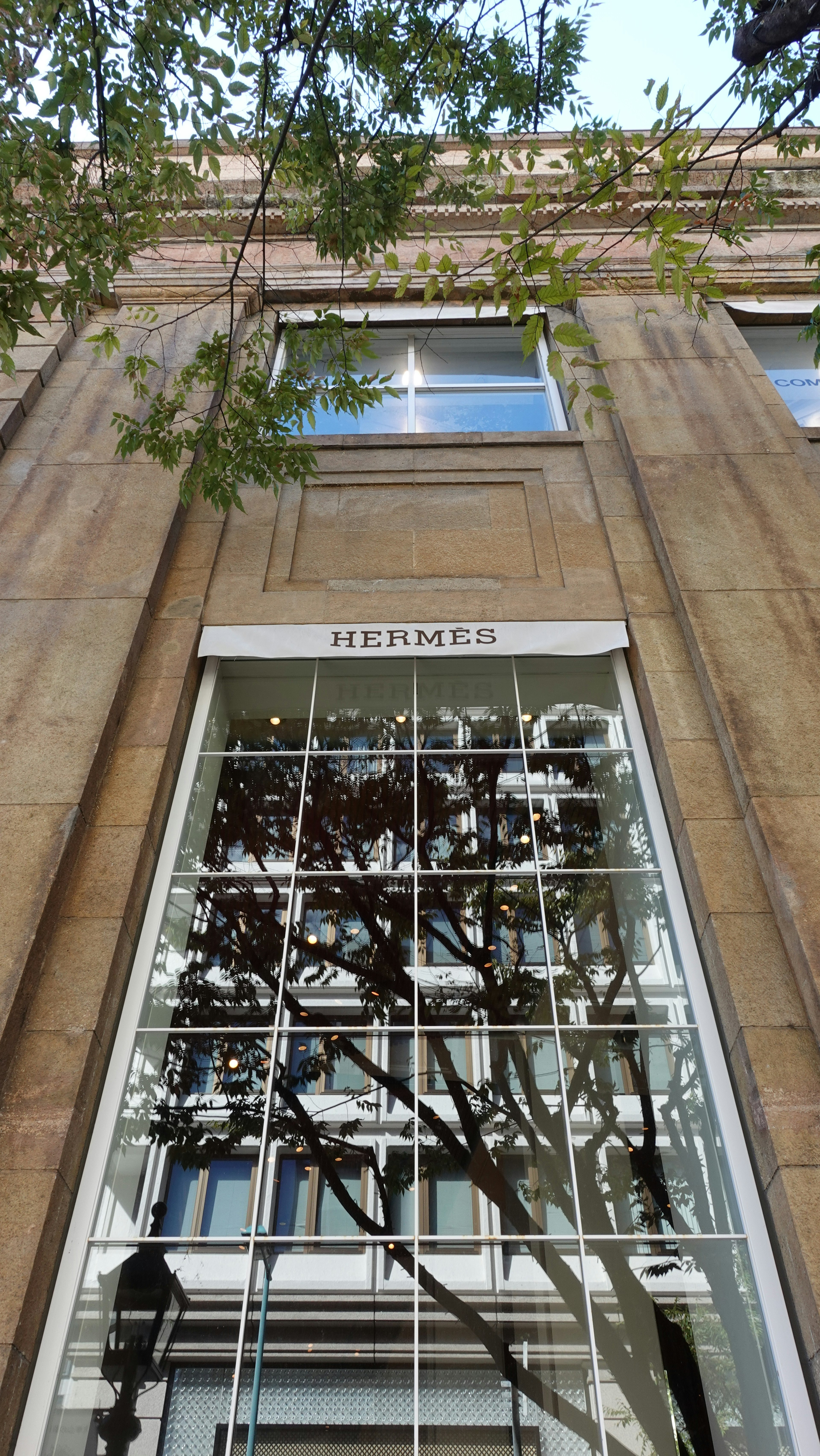 Imagen del exterior de la tienda Hermès con grandes ventanas de vidrio y reflejos de árboles