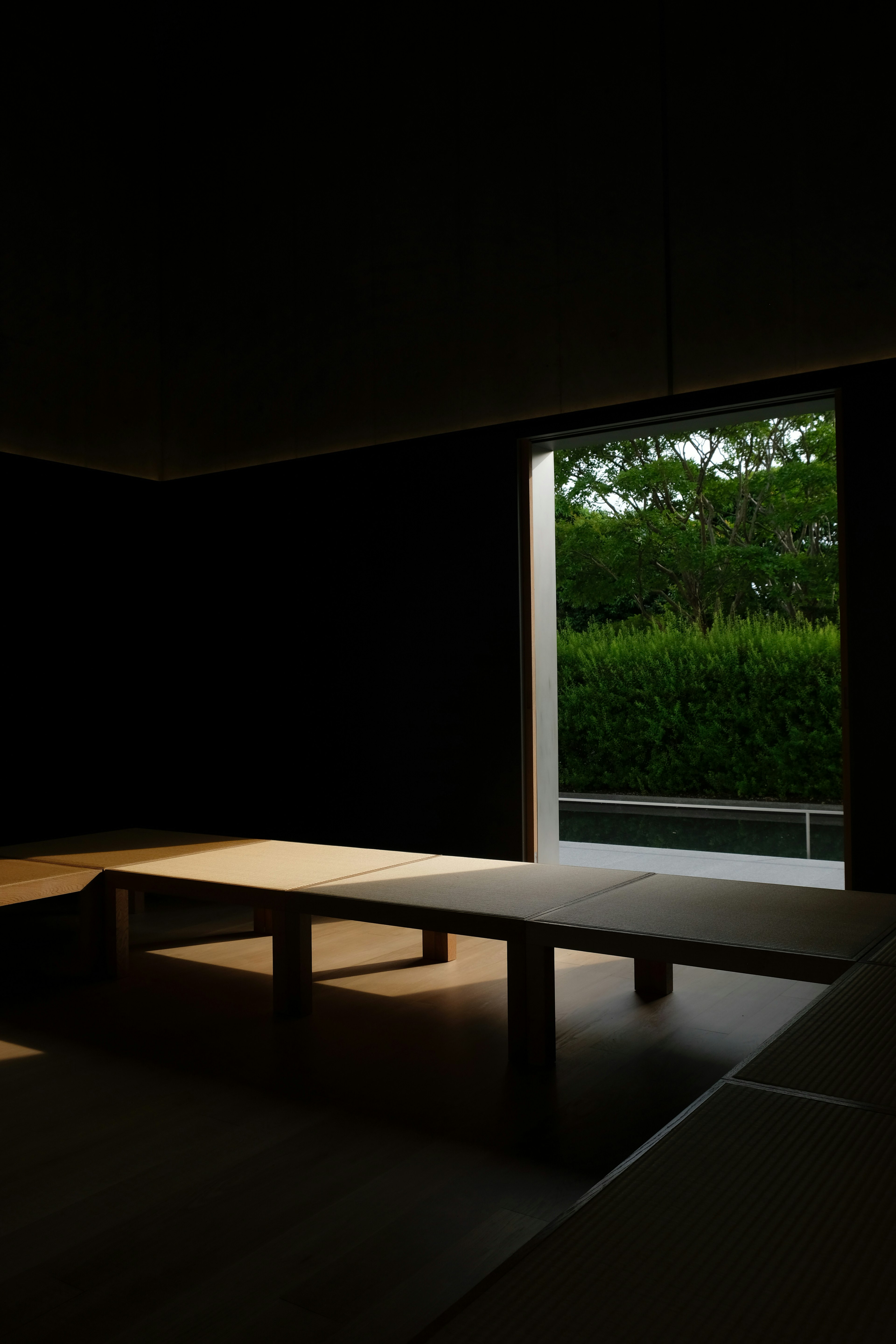 Un intérieur sombre avec une fenêtre lumineuse montrant de la verdure à l'extérieur et une table simple