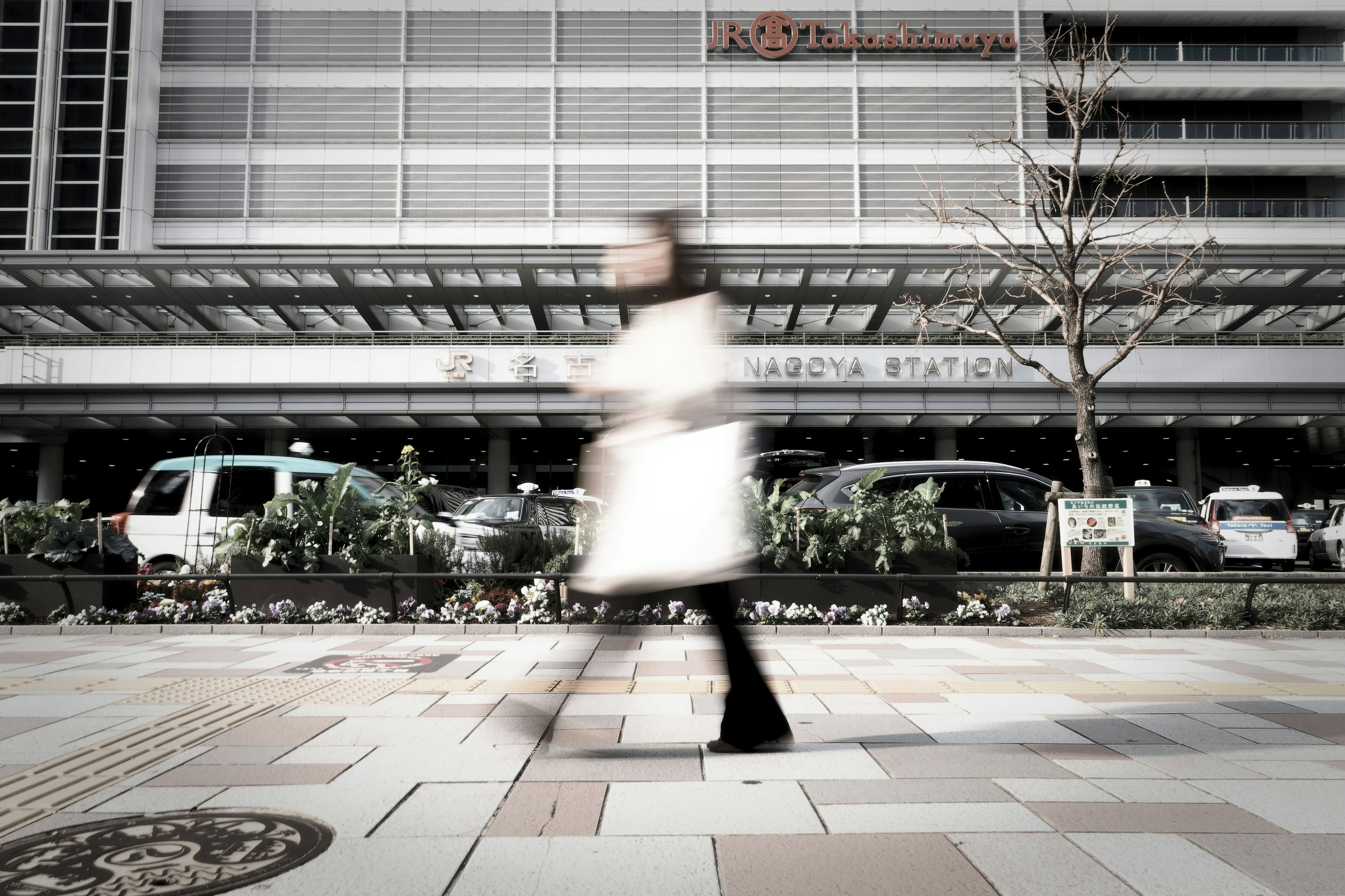 Una persona in un cappotto bianco che cammina in un ambiente urbano con un'insegna della stazione e veicoli sullo sfondo