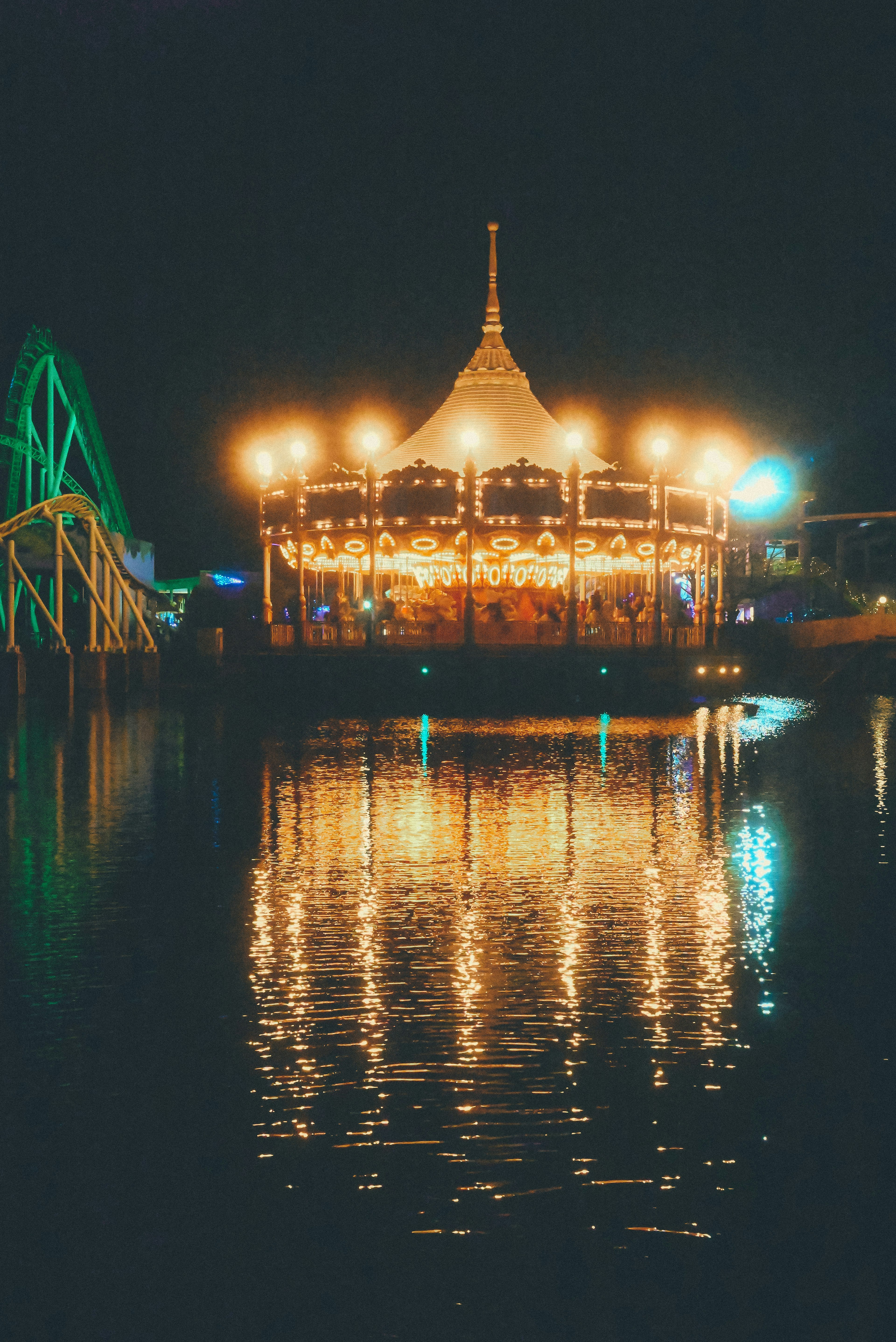 Bright carousel illuminated at night reflecting on water with colorful rides in the background