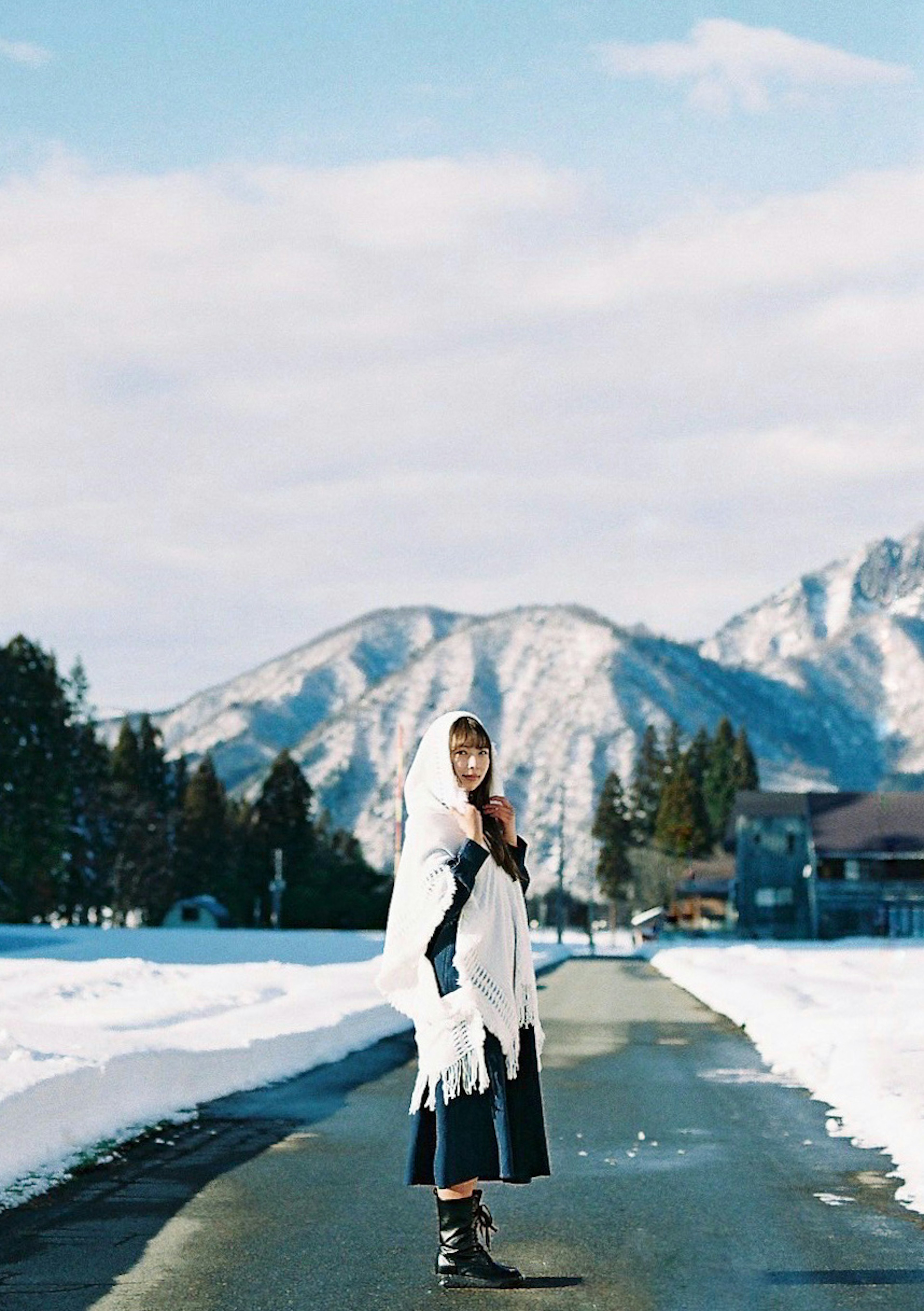 一位穿著白色披肩的女性在雪地小路上走，背景是山脈