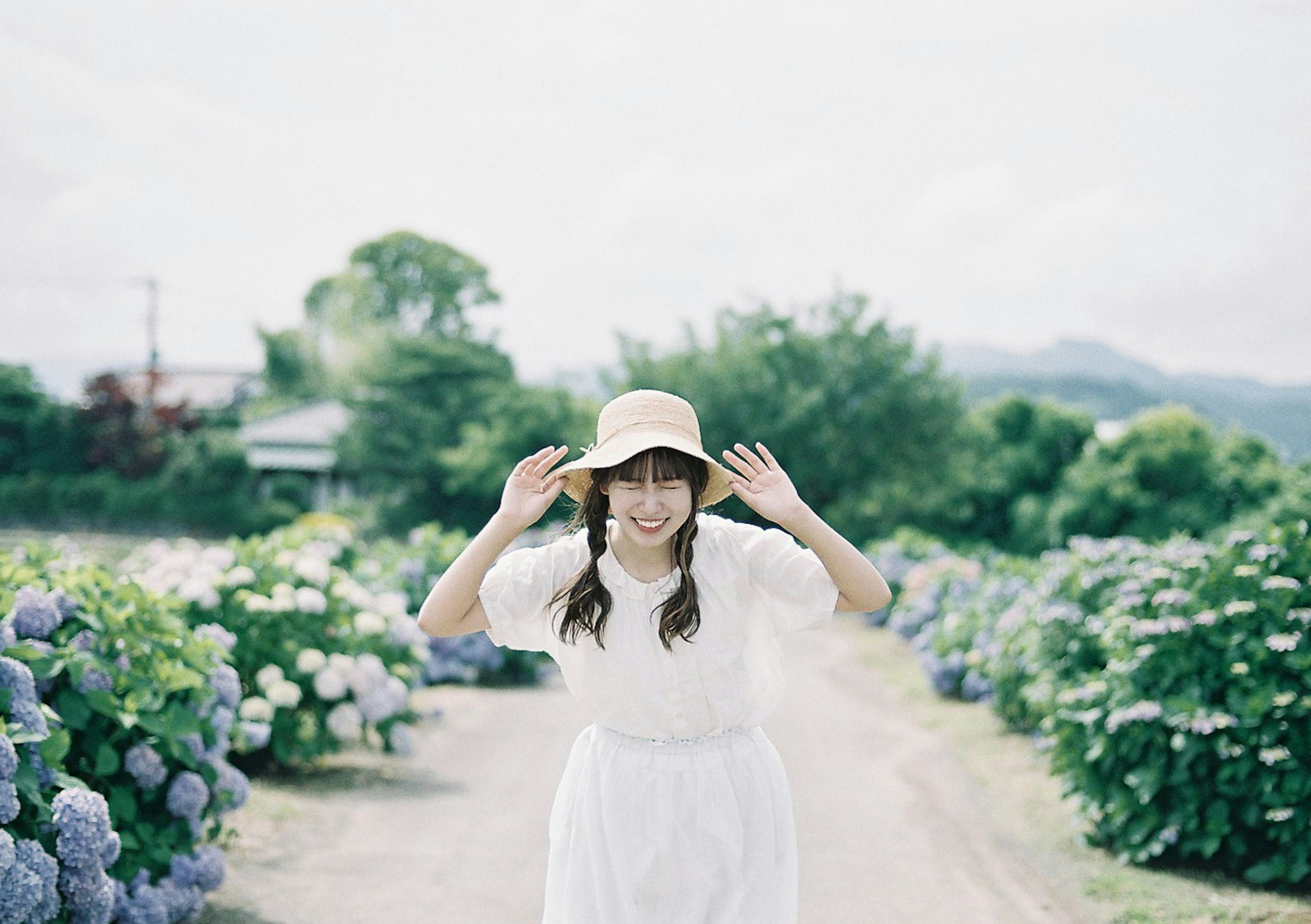 Una mujer con un vestido blanco y sombrero sonríe y posa rodeada de hortensias azules